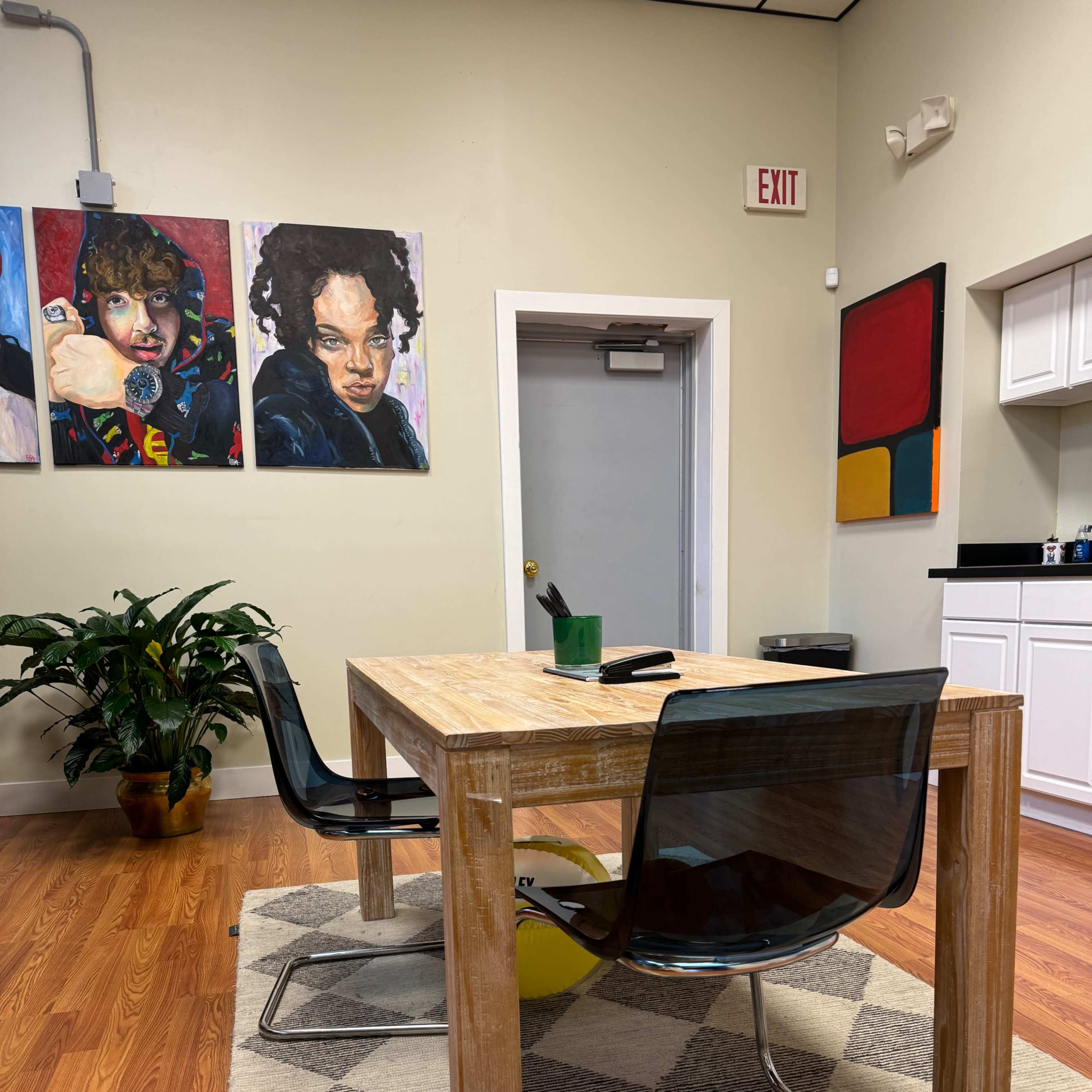 A wooden table with transparent chairs is set in an office space featuring colorful artwork on the walls and a plant in the corner.