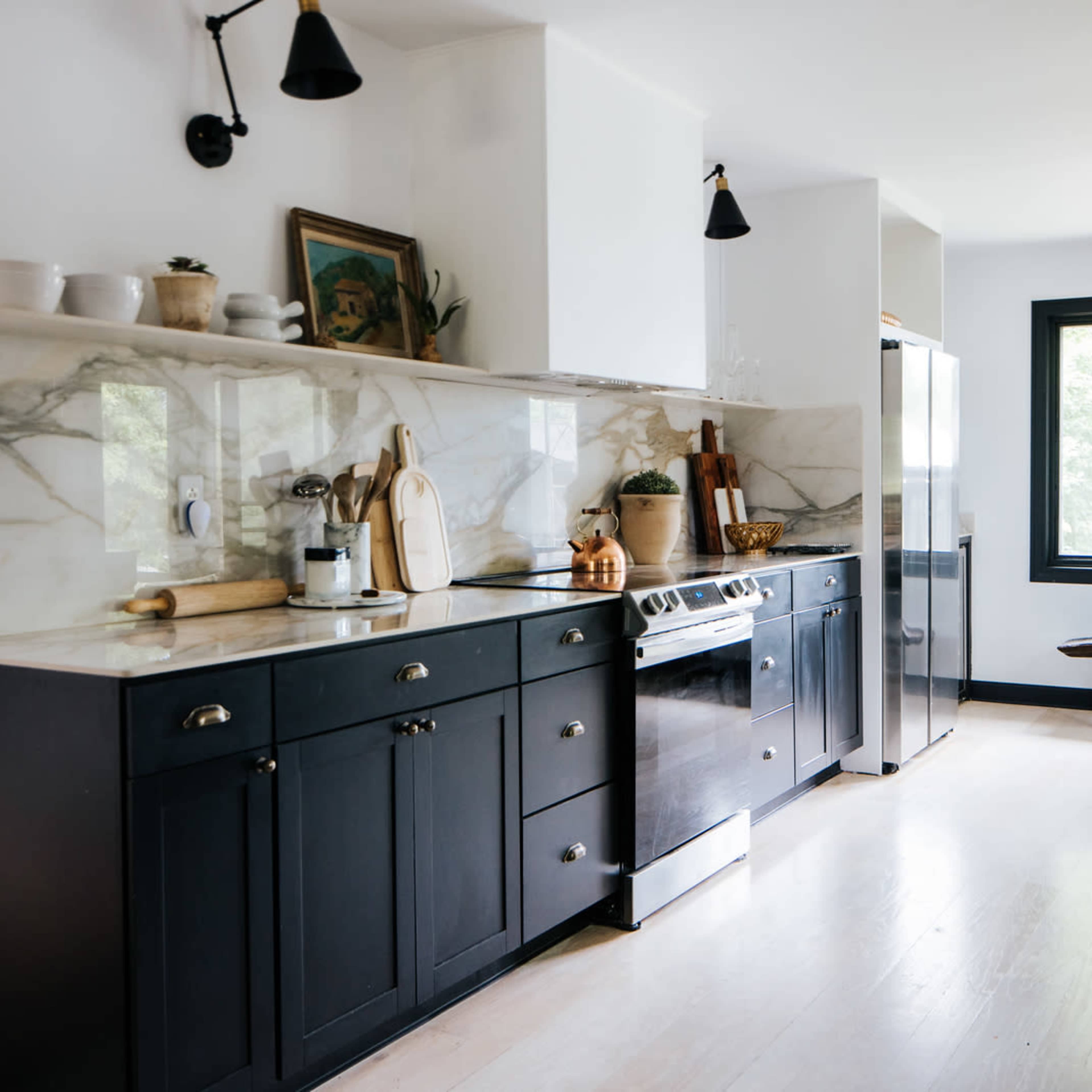 A modern kitchen features black cabinetry, marble countertops, stainless steel appliances, and a wooden bench near a window.