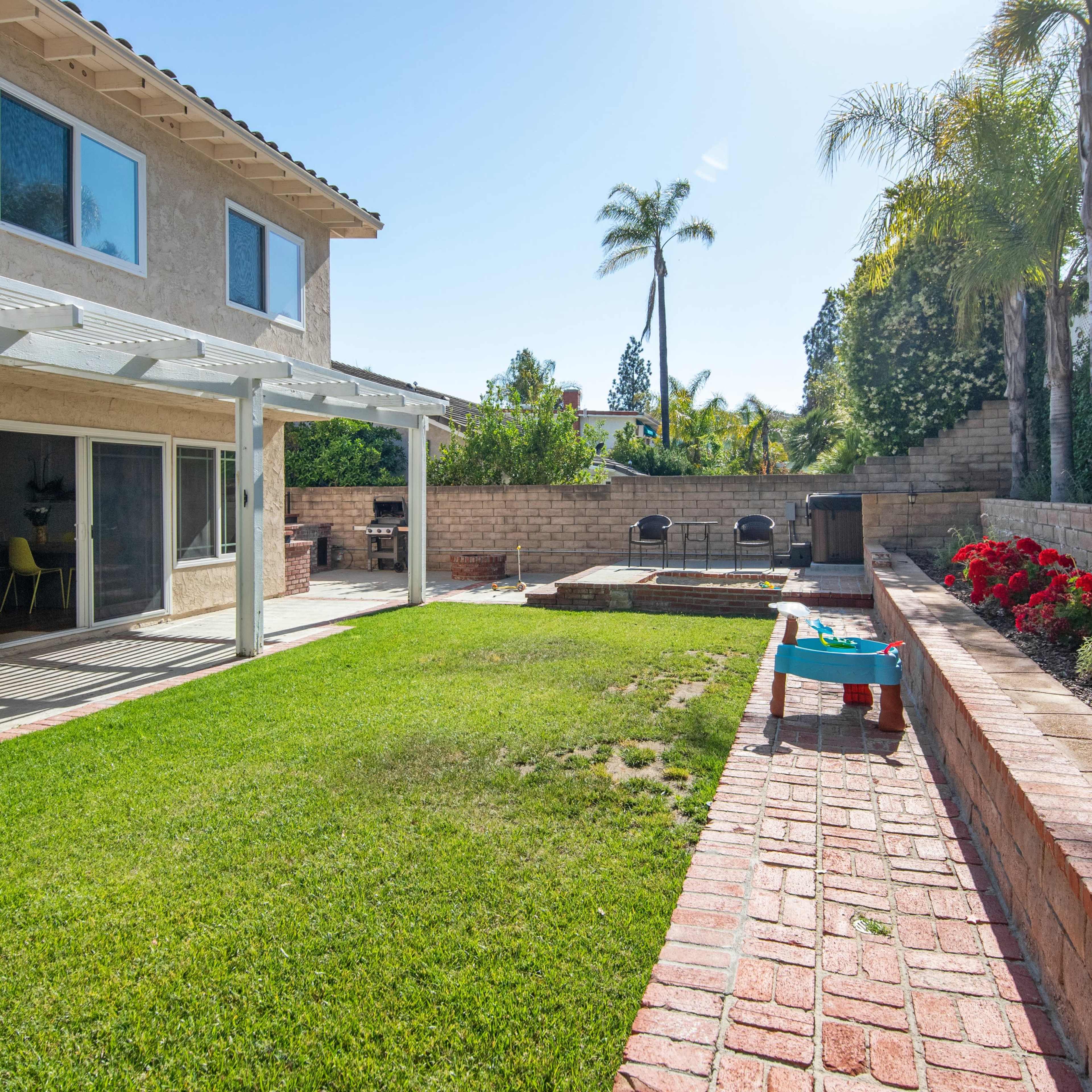 A backyard with a green lawn, brick pathways, a patio with chairs, and a small pool surrounded by landscaped garden beds.