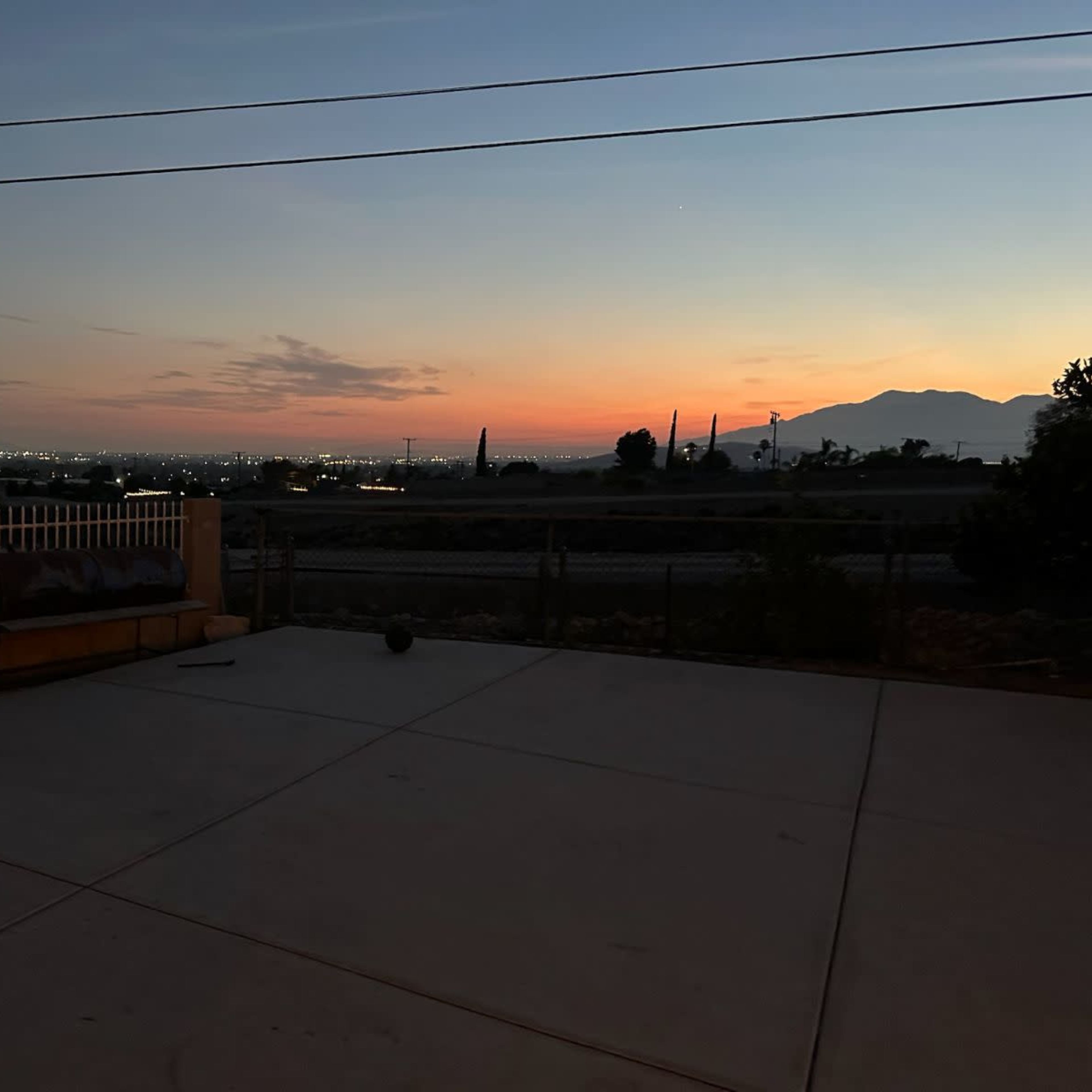 A concrete patio overlooking a cityscape at dusk, with mountains in the background and a horizon painted in shades of orange and blue.