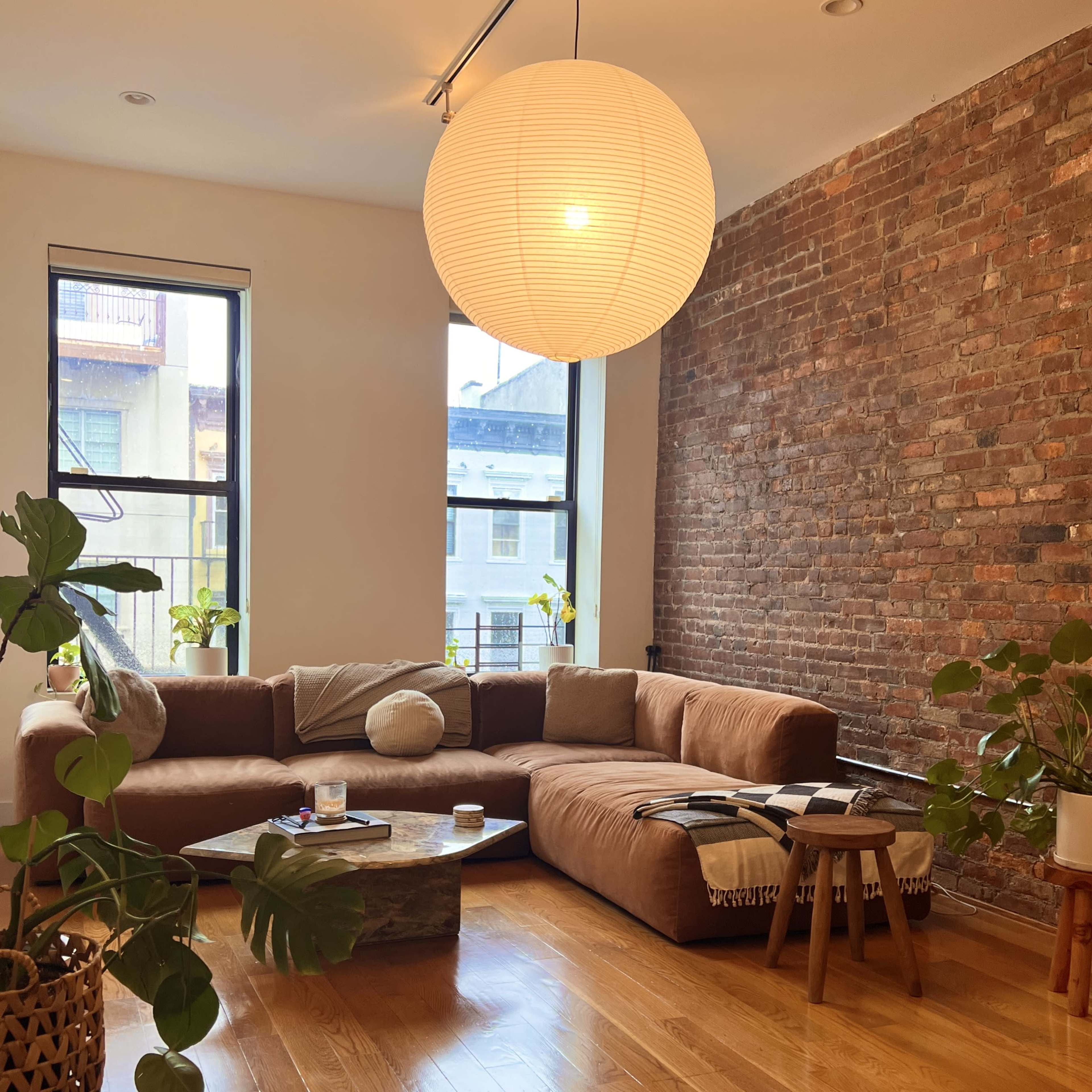 A living room with a brown sectional sofa, a round coffee table, a large paper lantern light fixture, and several plants against a brick wall.