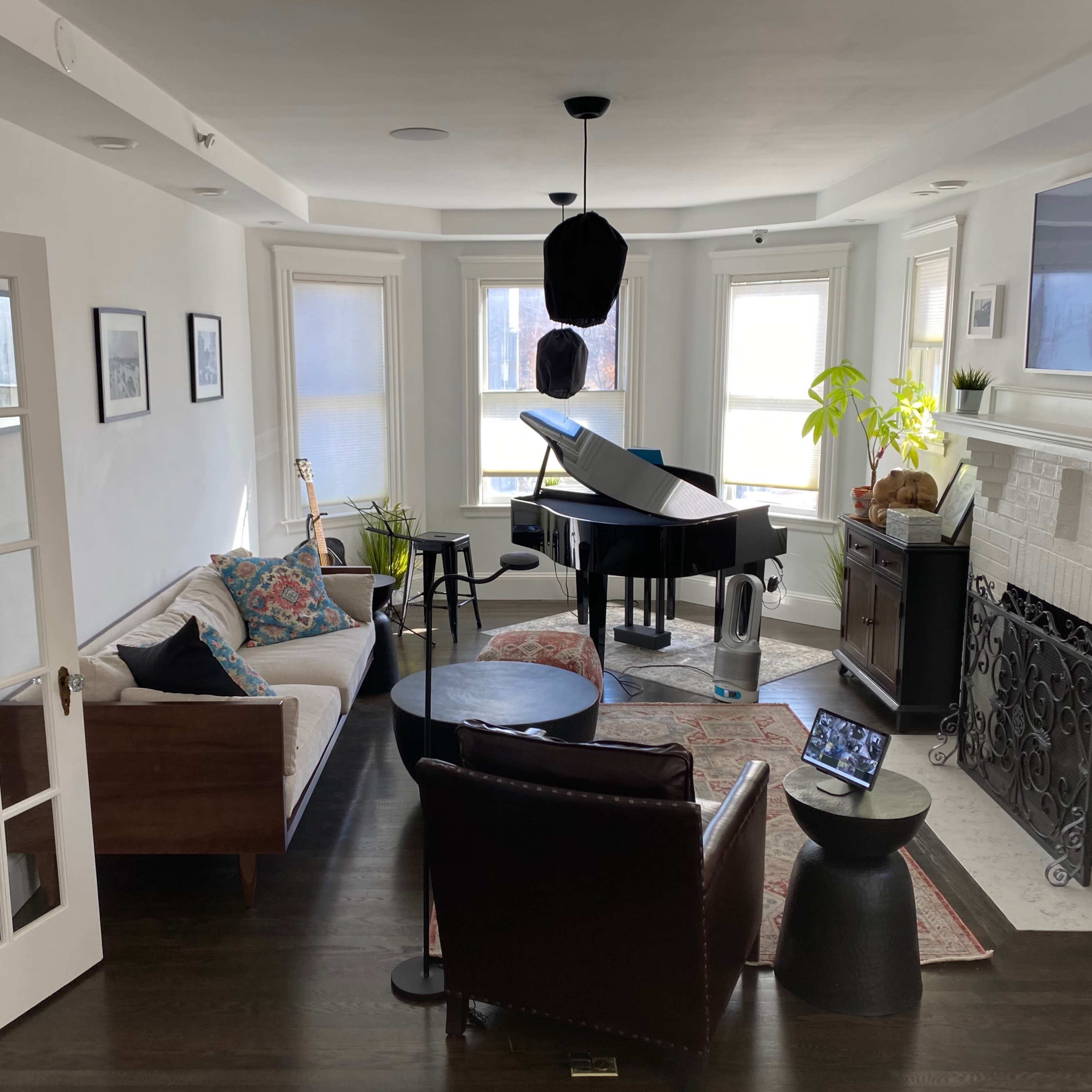 The scene shows a bright living room featuring a grand piano, a couch, a round table, and a fireplace, all arranged around large windows.