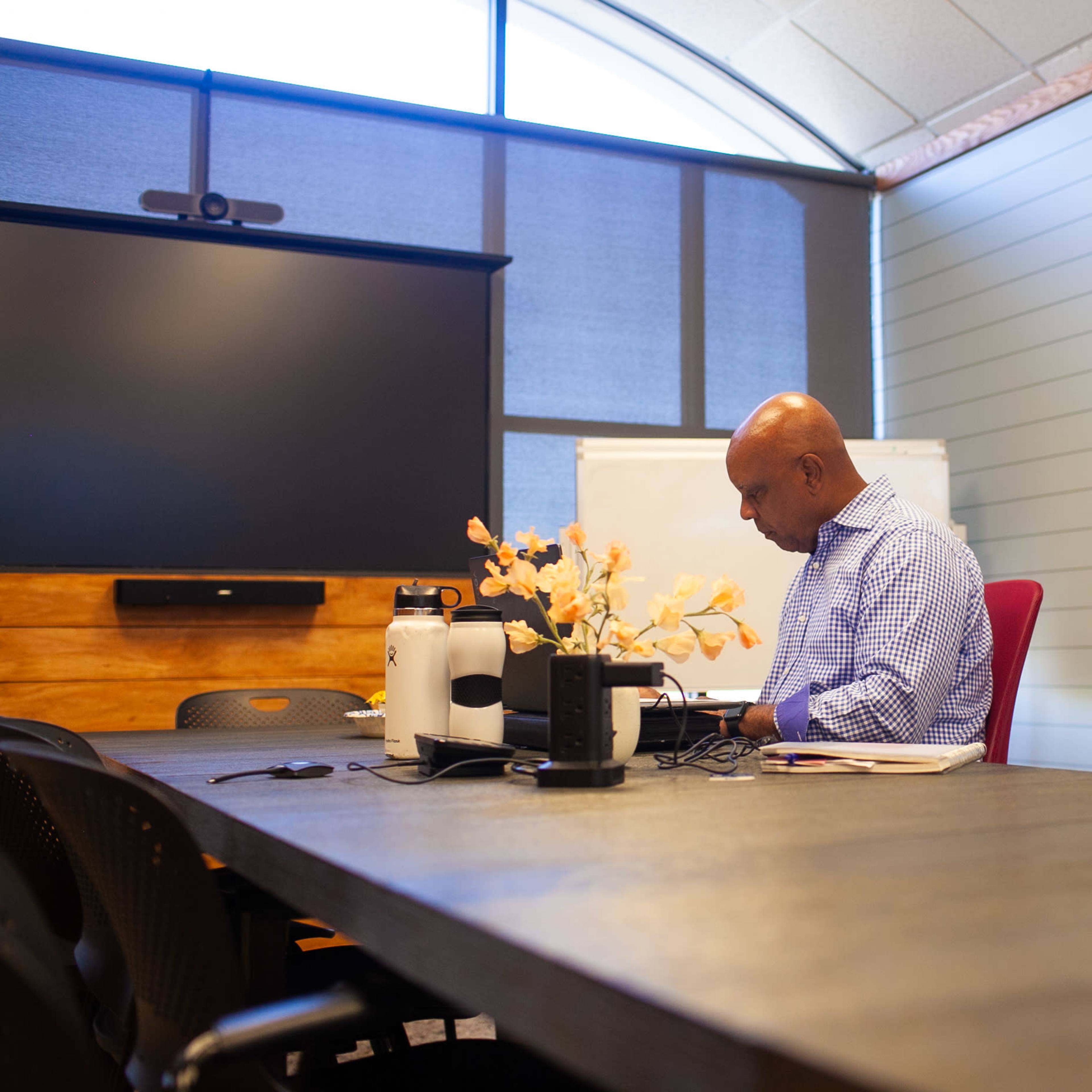 A man is seated at a conference table in a meeting room, working on a laptop with a floral arrangement in the center.
