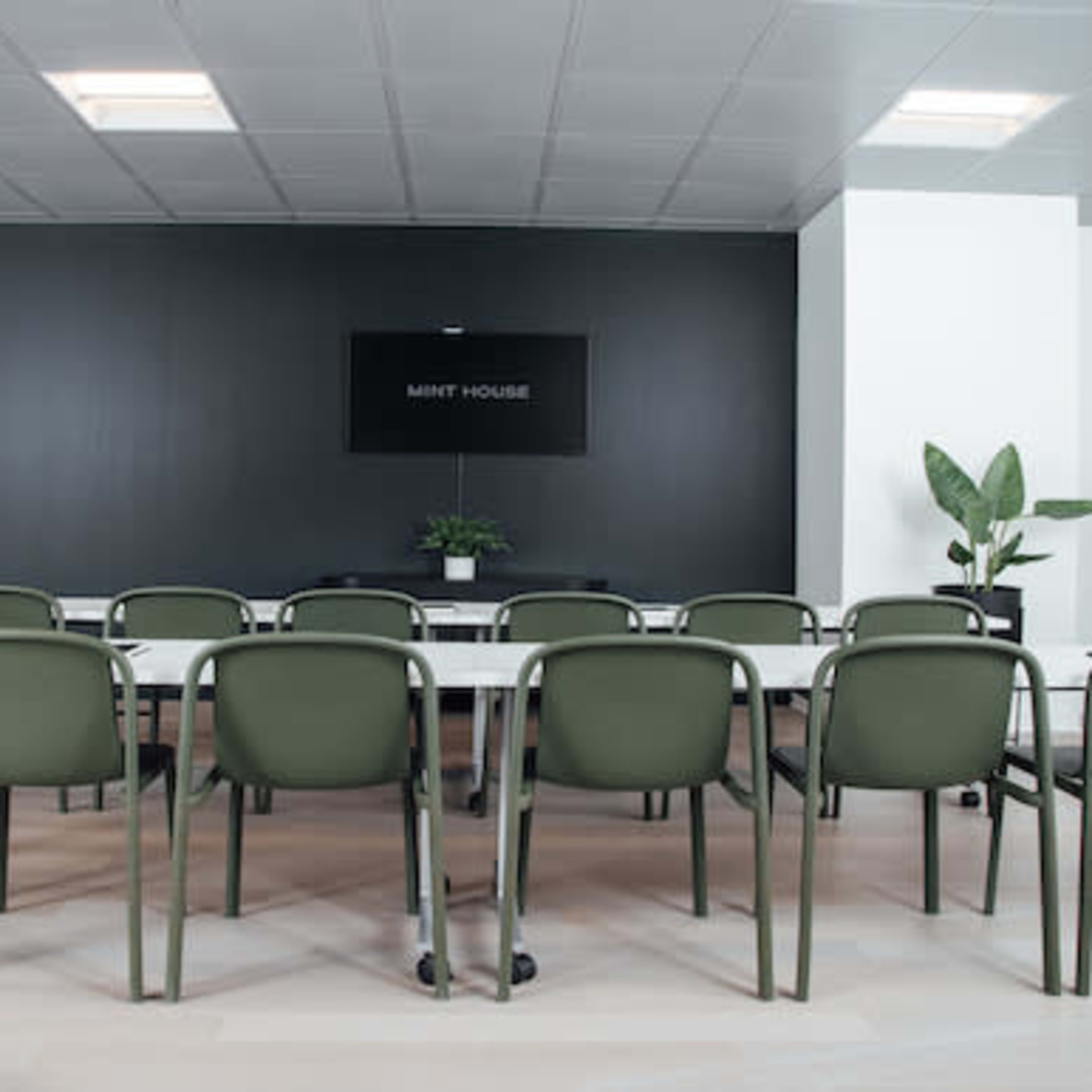 A modern conference room features a long table surrounded by green chairs, with a black wall that includes a sign labeled "MINT HOUSE."