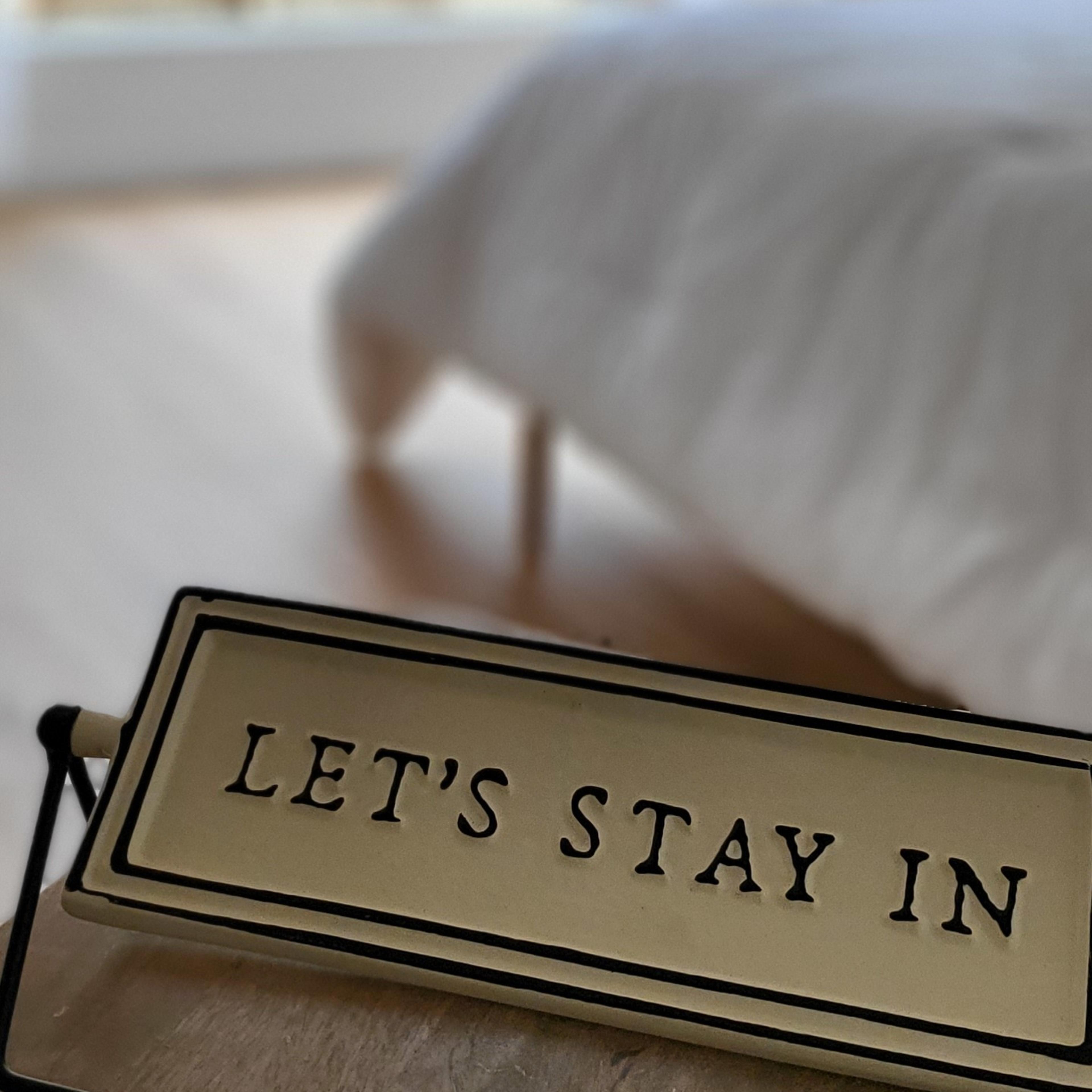 A sign that reads "LET'S STAY IN" is displayed on a table in a bedroom with a window showing a green outdoor view.