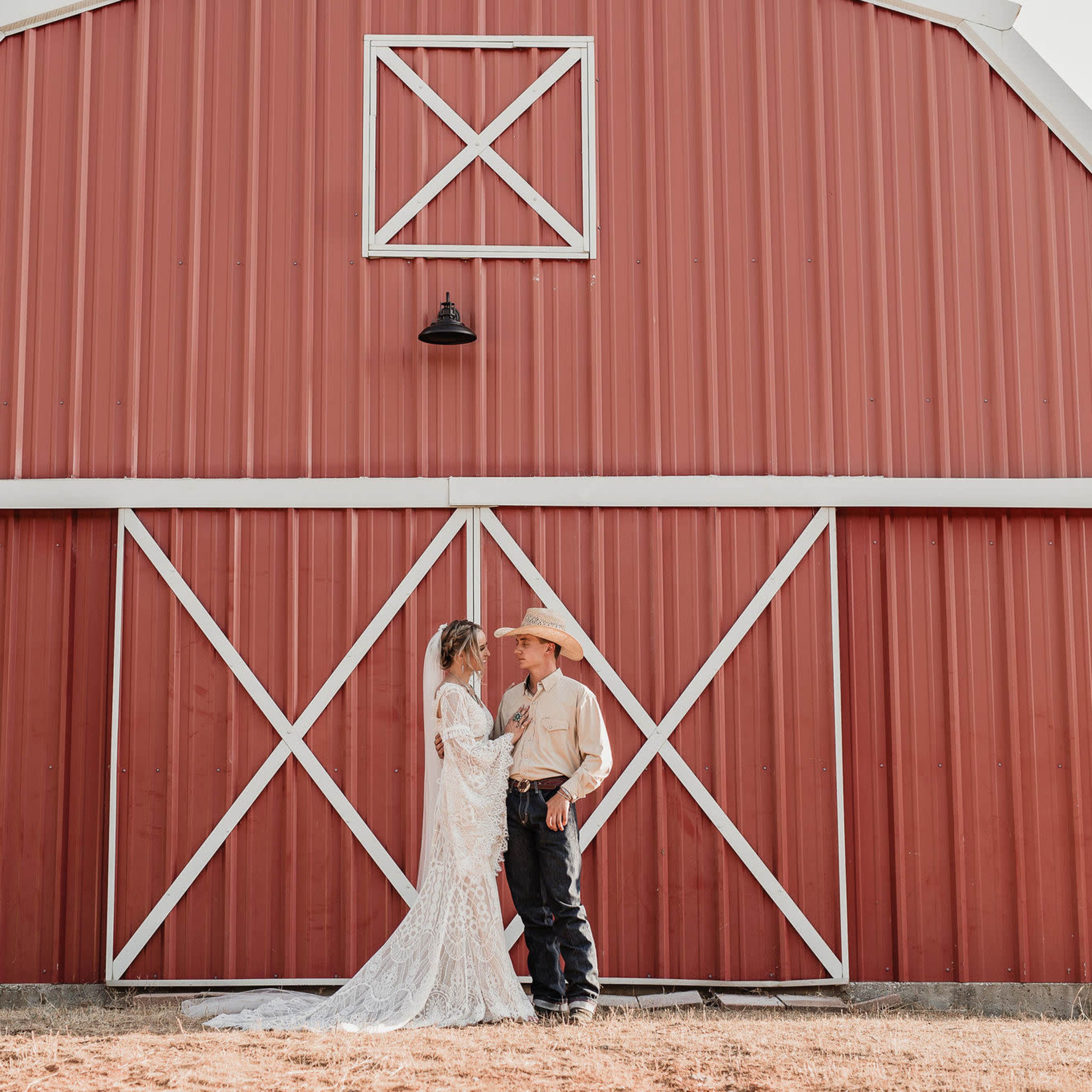 A couple stands closely together in front of a red barn, with the bride in a white dress and the groom in cowboy attire.