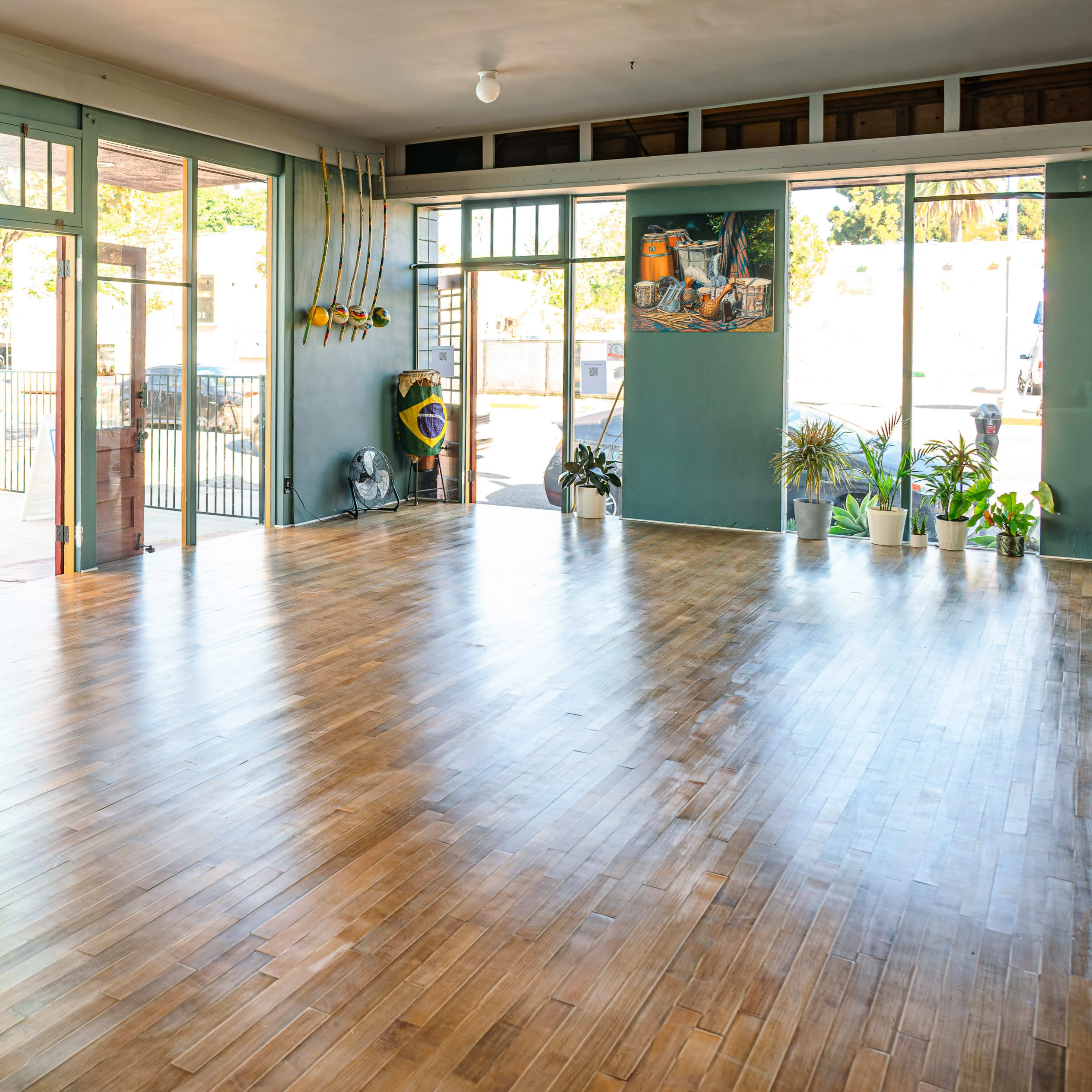 A spacious, empty room with wooden floors, large windows, and potted plants along the walls.