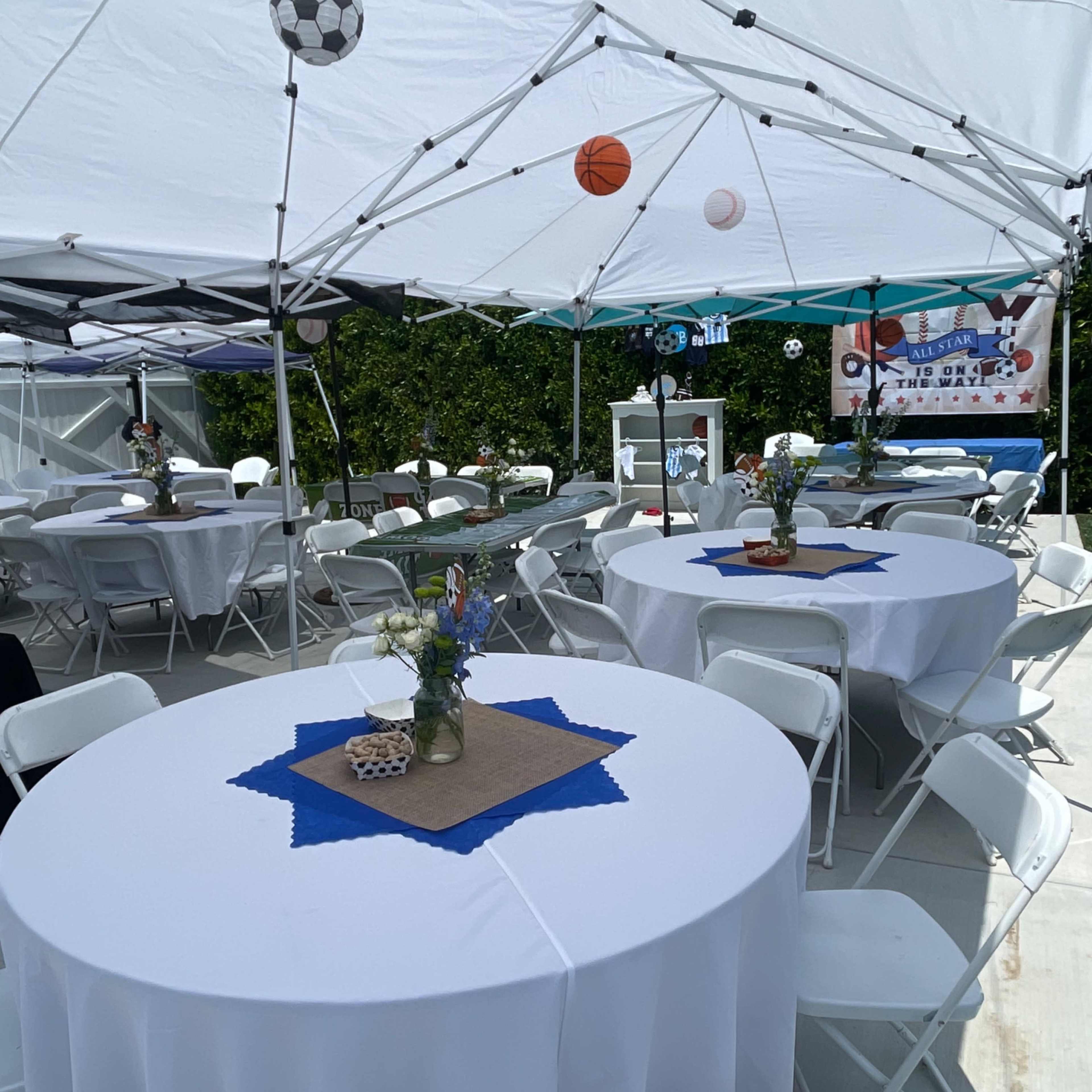 A tent set up for a sports-themed party features several tables with white tablecloths, decorated with centerpieces, and sports balls hanging above.