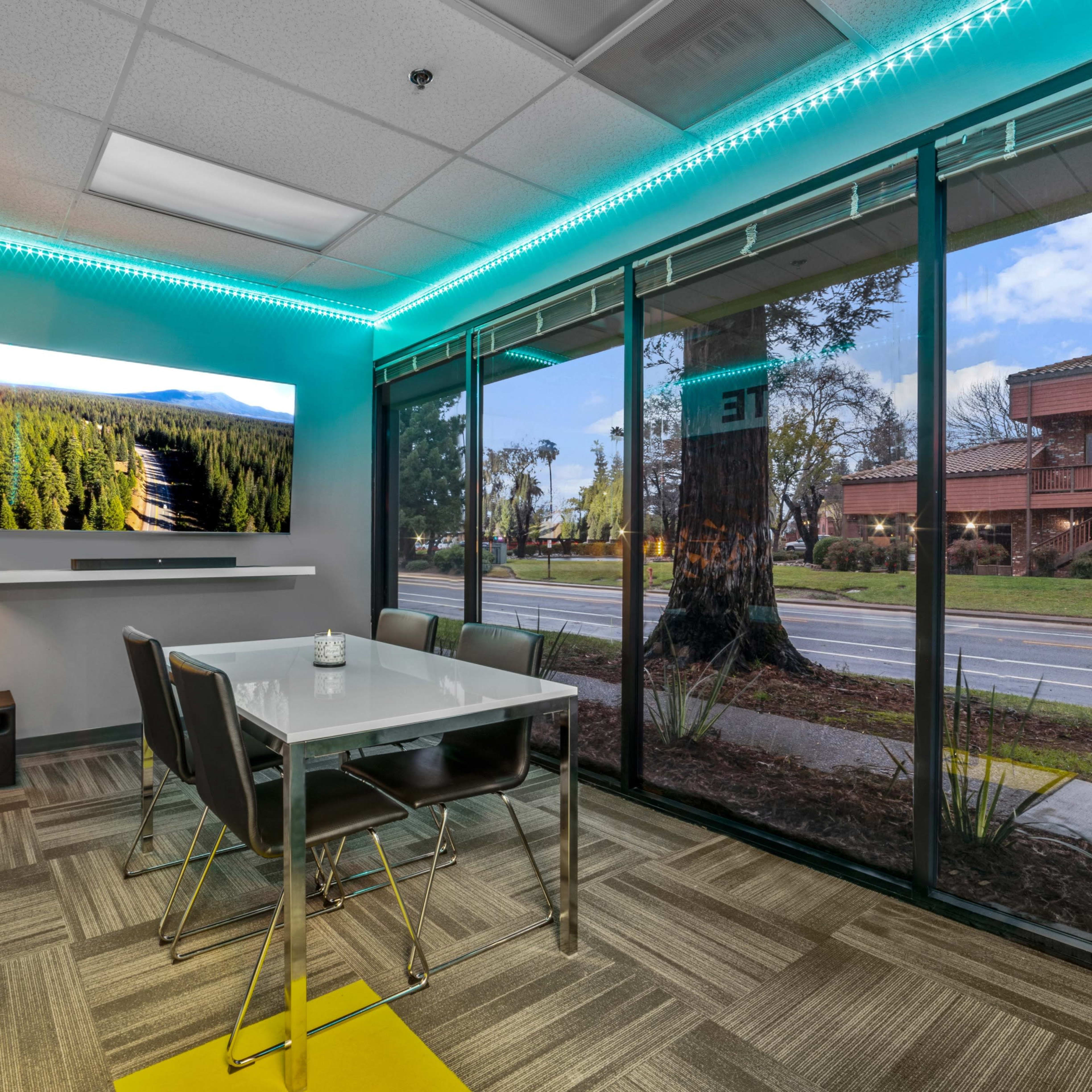 A modern conference room features a glass wall with a view of the street, a large screen displaying a nature scene, and a table surrounded by chairs.