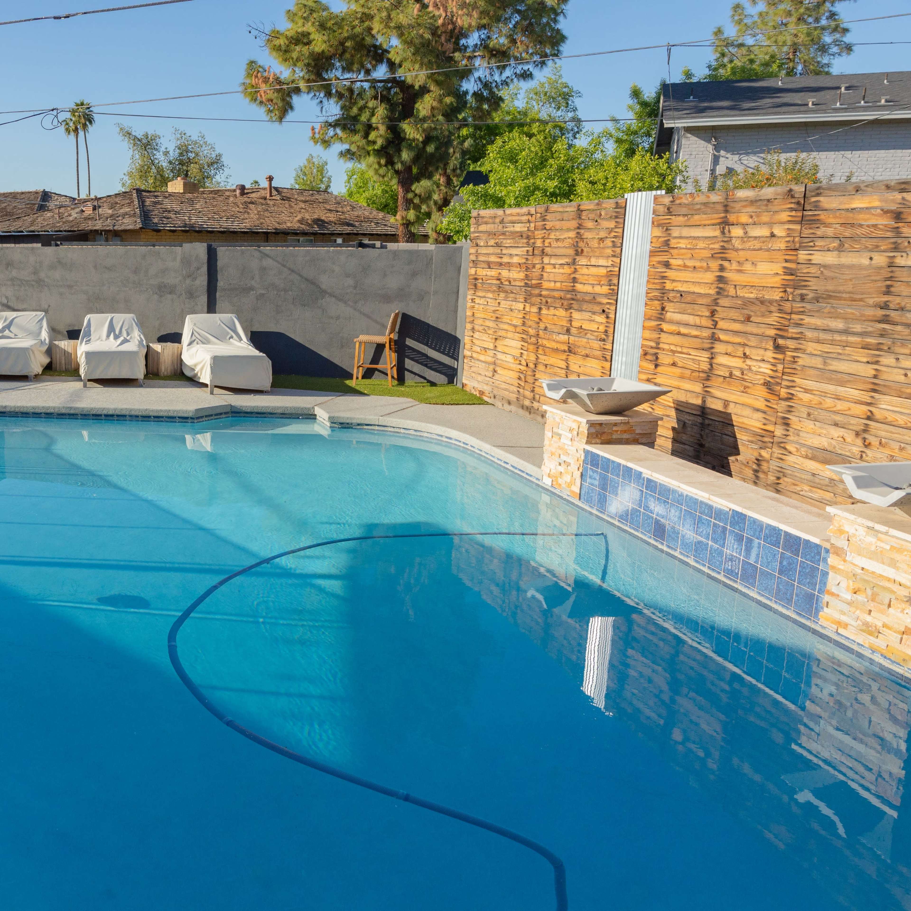 A clear swimming pool is surrounded by a fenced area with lounge chairs and a wooden fence in the background.