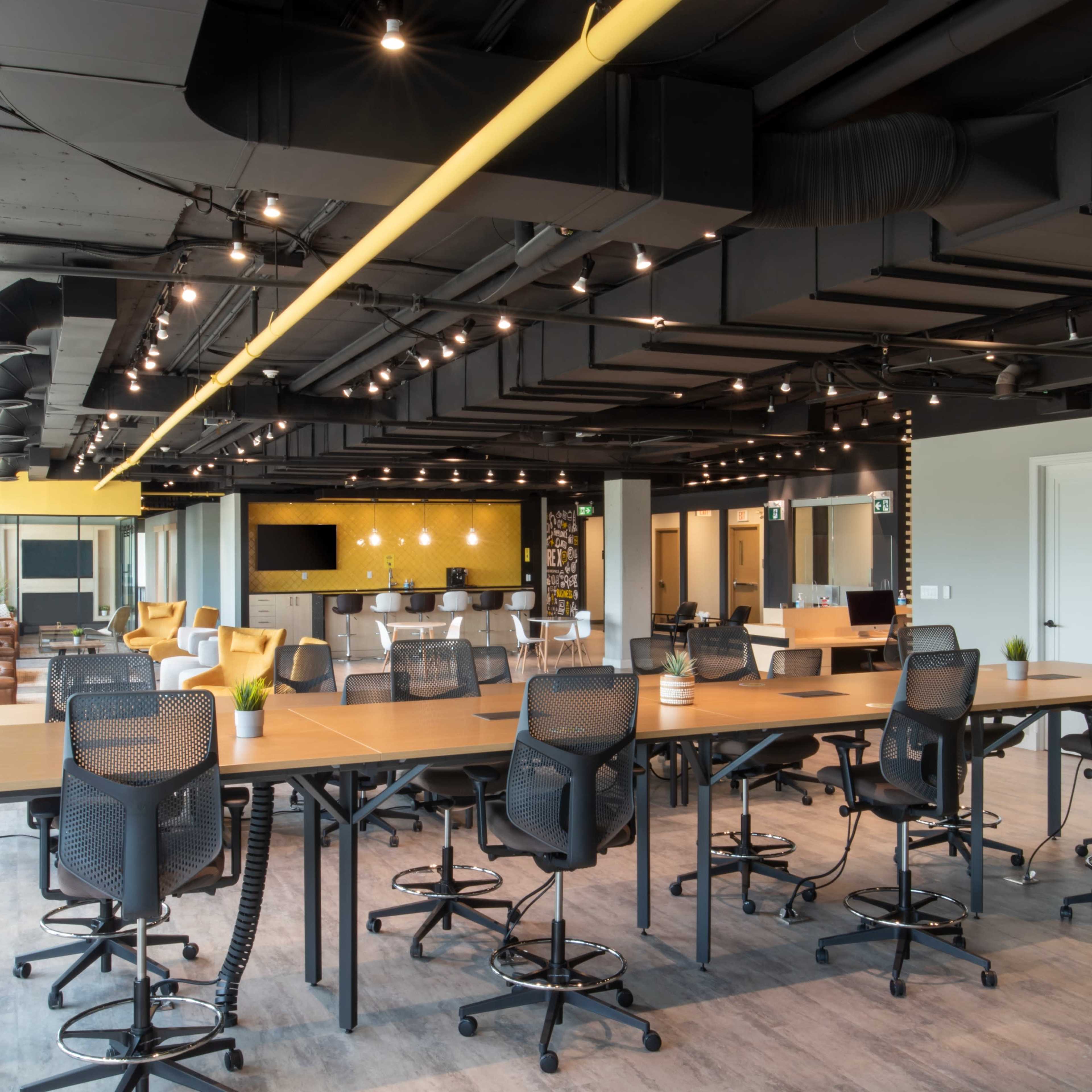 A modern office space with a large communal table surrounded by tall chairs, features bright overhead lighting, and various seating arrangements in the background.
