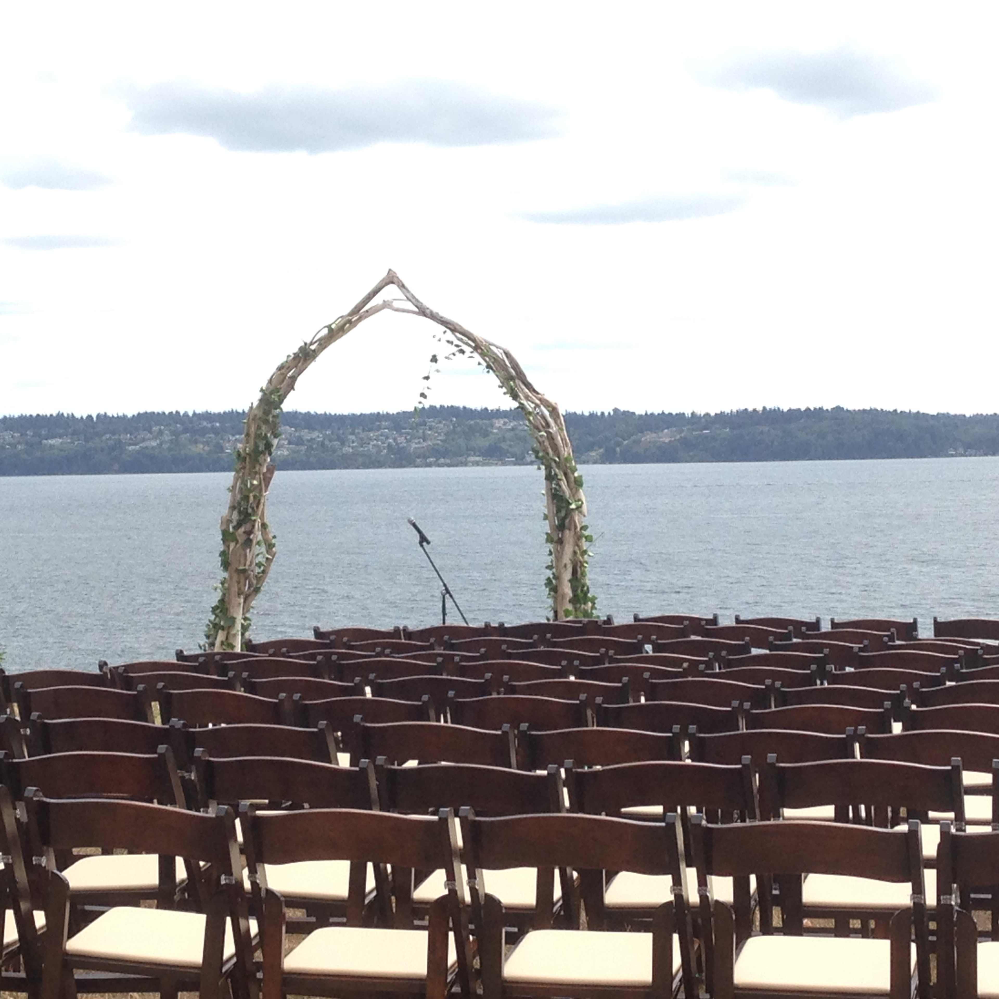 A wedding altar made of wooden branches is set up by a lake, with rows of empty chairs facing it.
