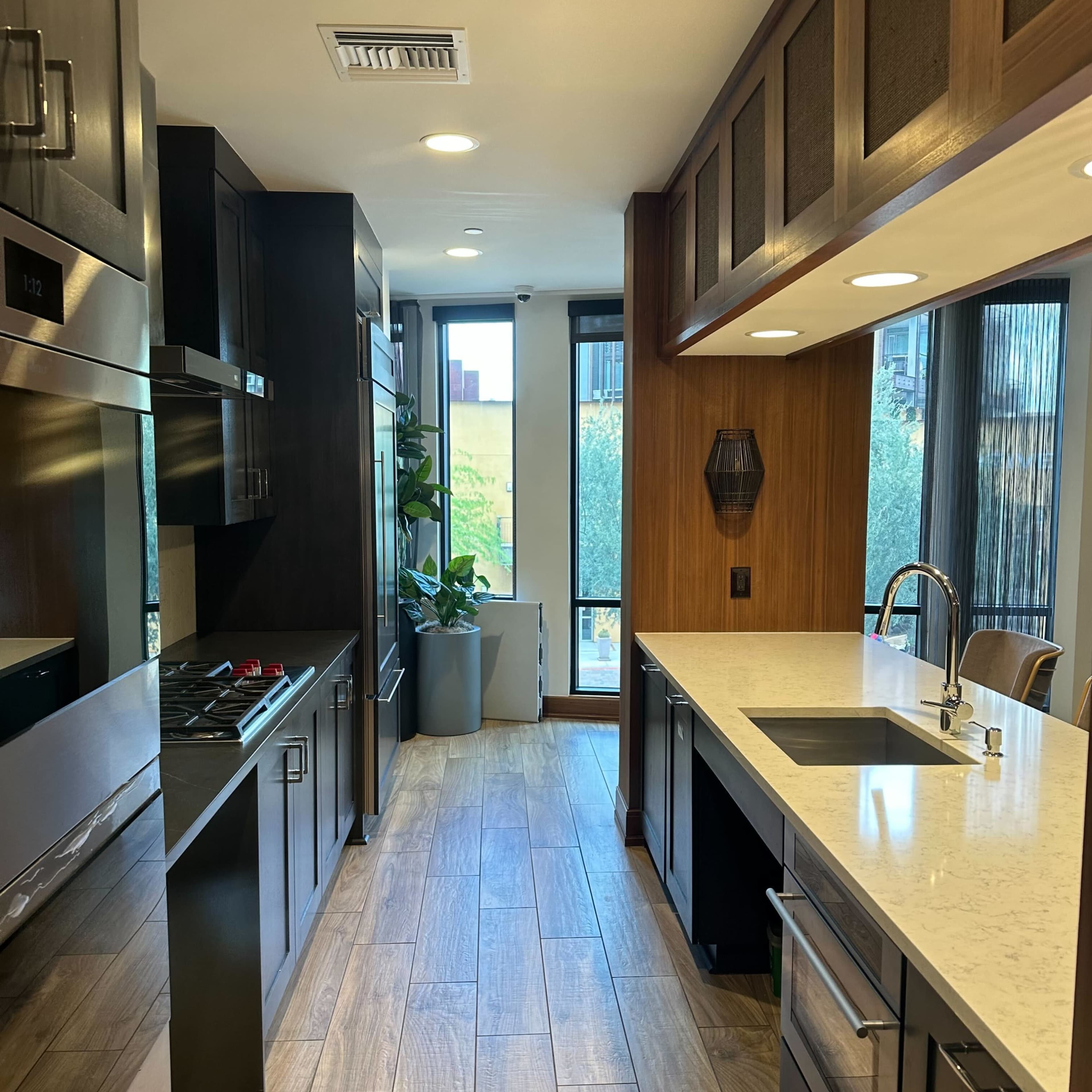 The kitchen features sleek black cabinetry, stainless steel appliances, and a white countertop, with a view of greenery through large windows.