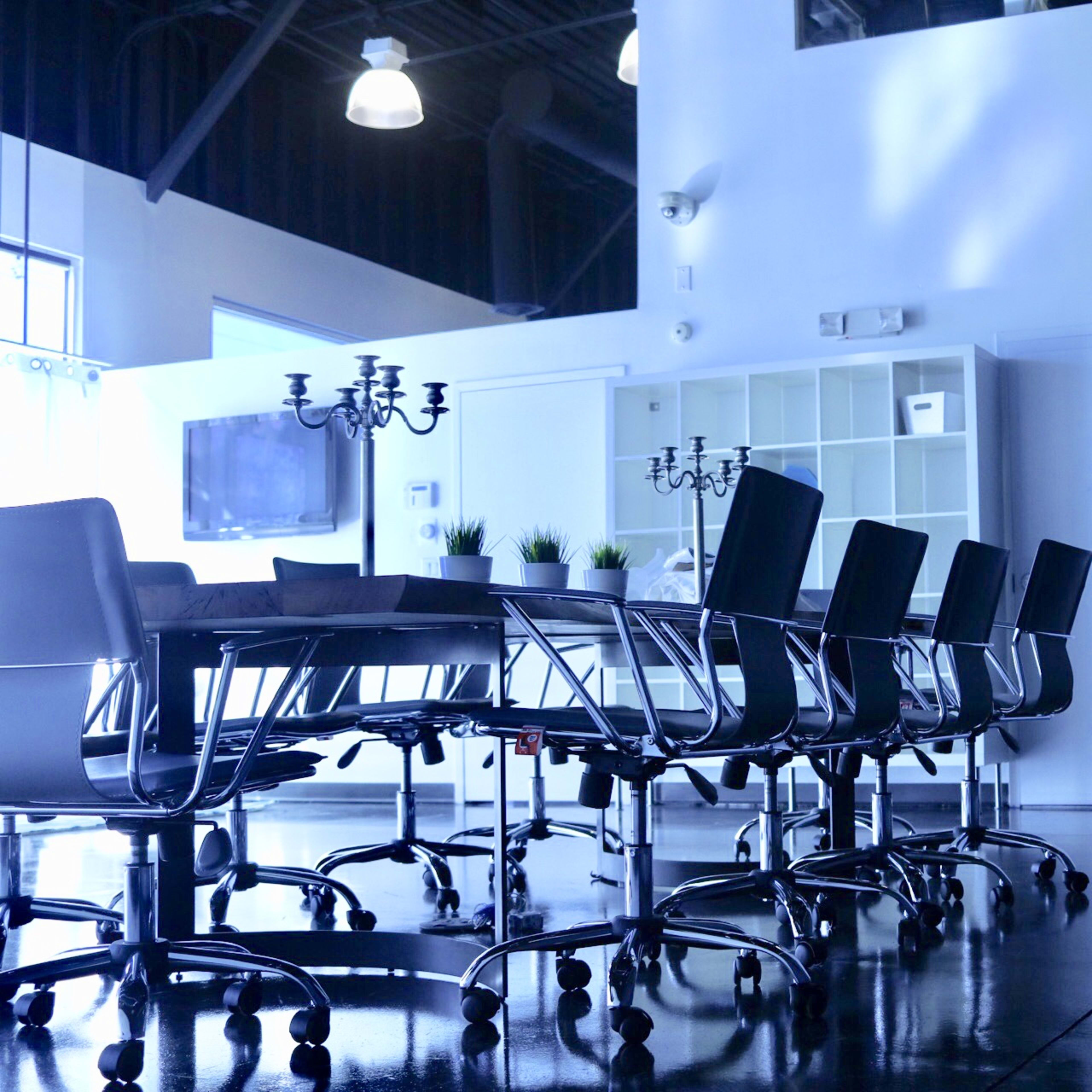 A long conference table with black chairs is placed in a bright, minimalist meeting room featuring a television and a shelf against the wall.