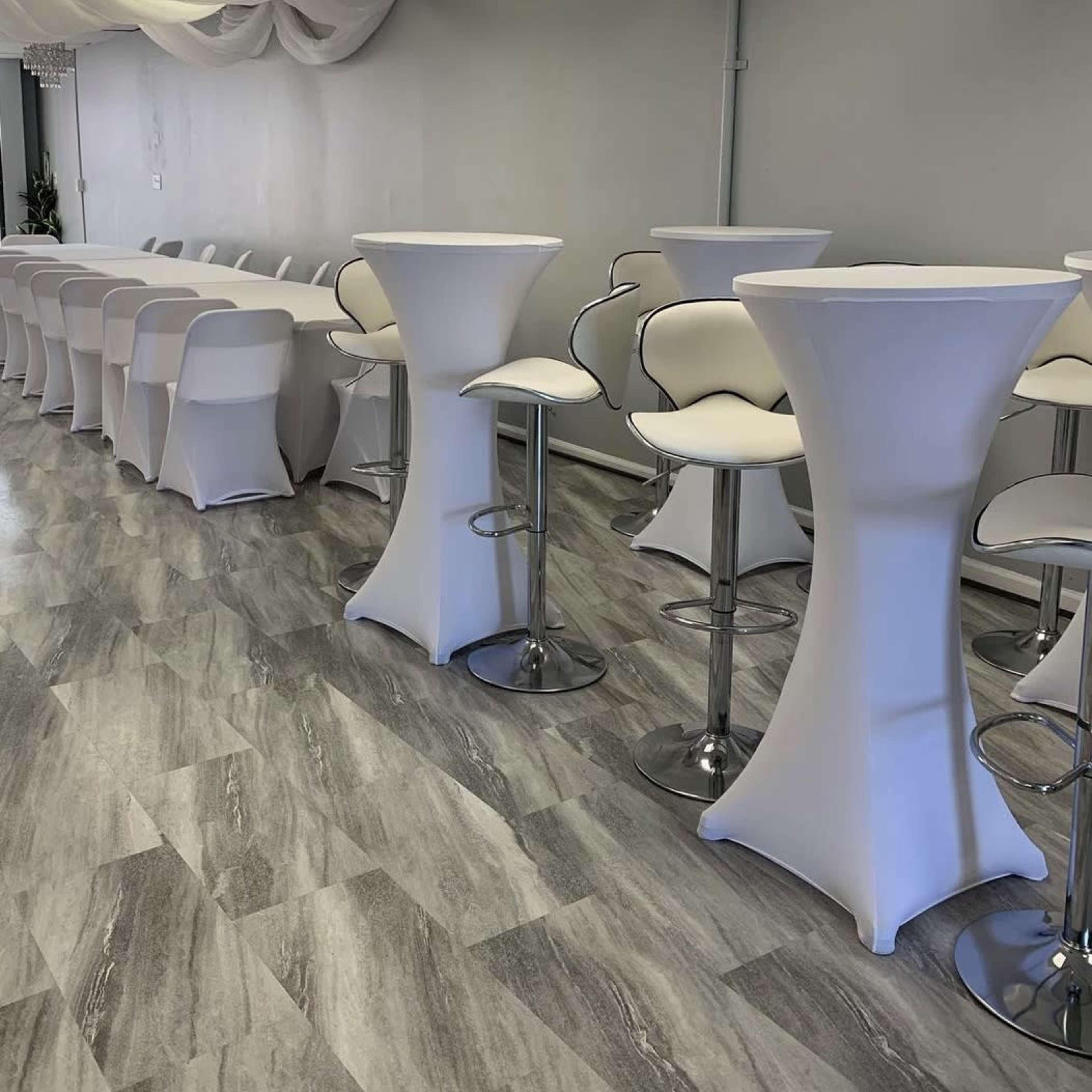 A spacious event room featuring white cocktail tables and high-backed stools arranged neatly along a light-colored wall.
