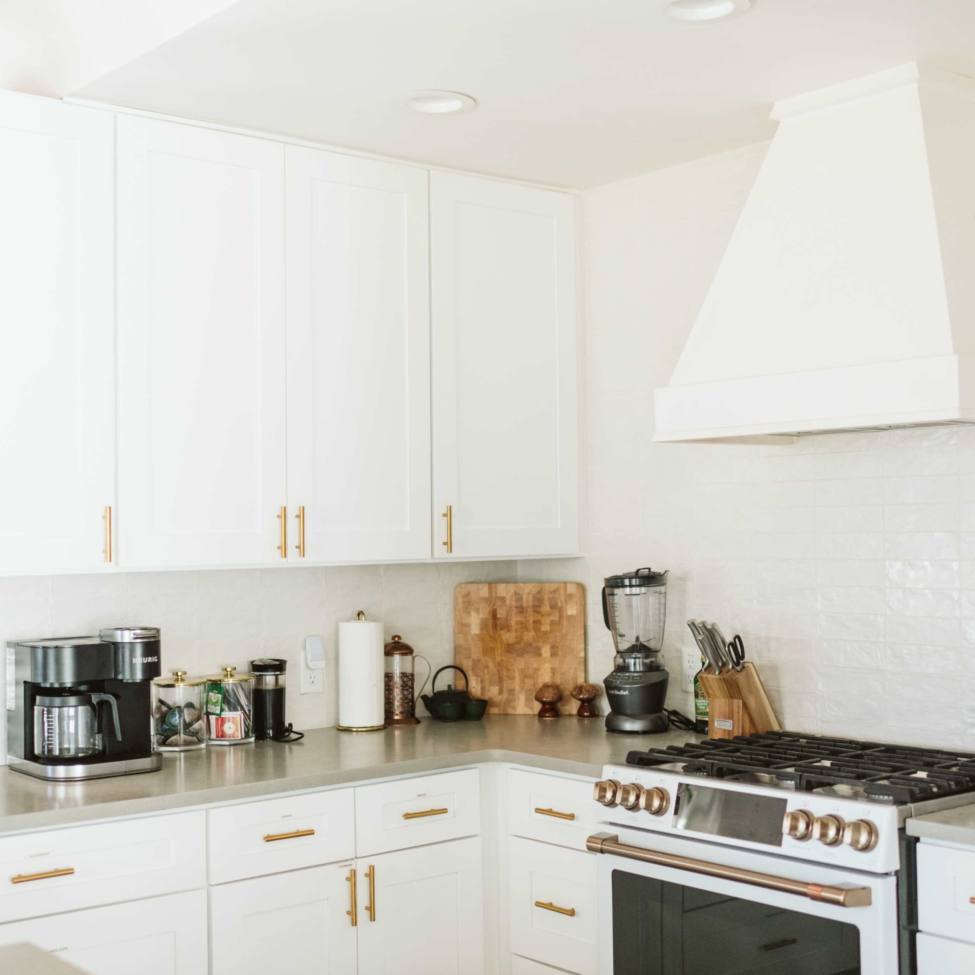 A bright kitchen features white cabinets with brass handles, a large hood above the stove, and various appliances like a coffee maker and blender on the countertop.