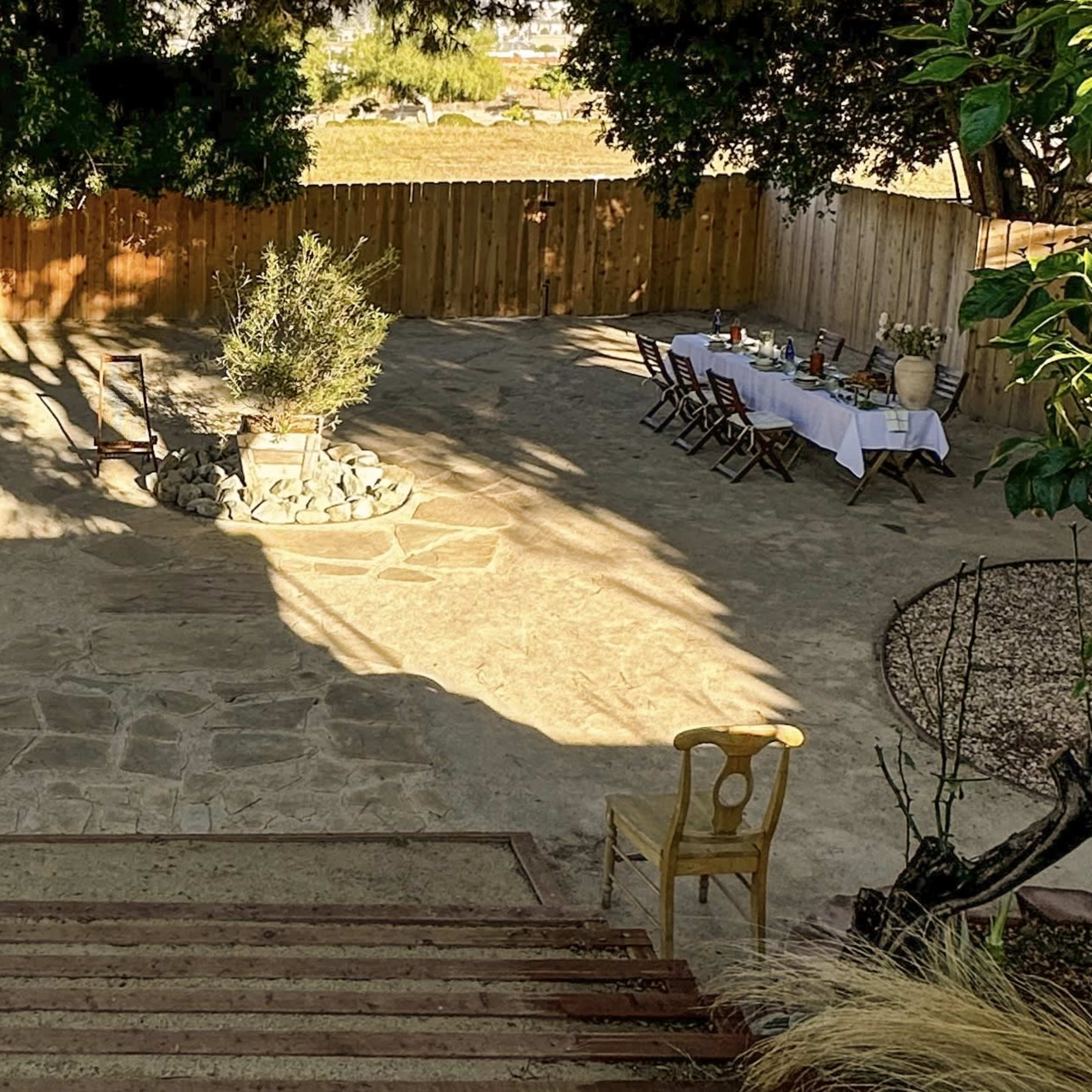 A spacious outdoor area with a stone pathway, a circular fire pit, several tables set for dining, and a wooden fence surrounding the scene.