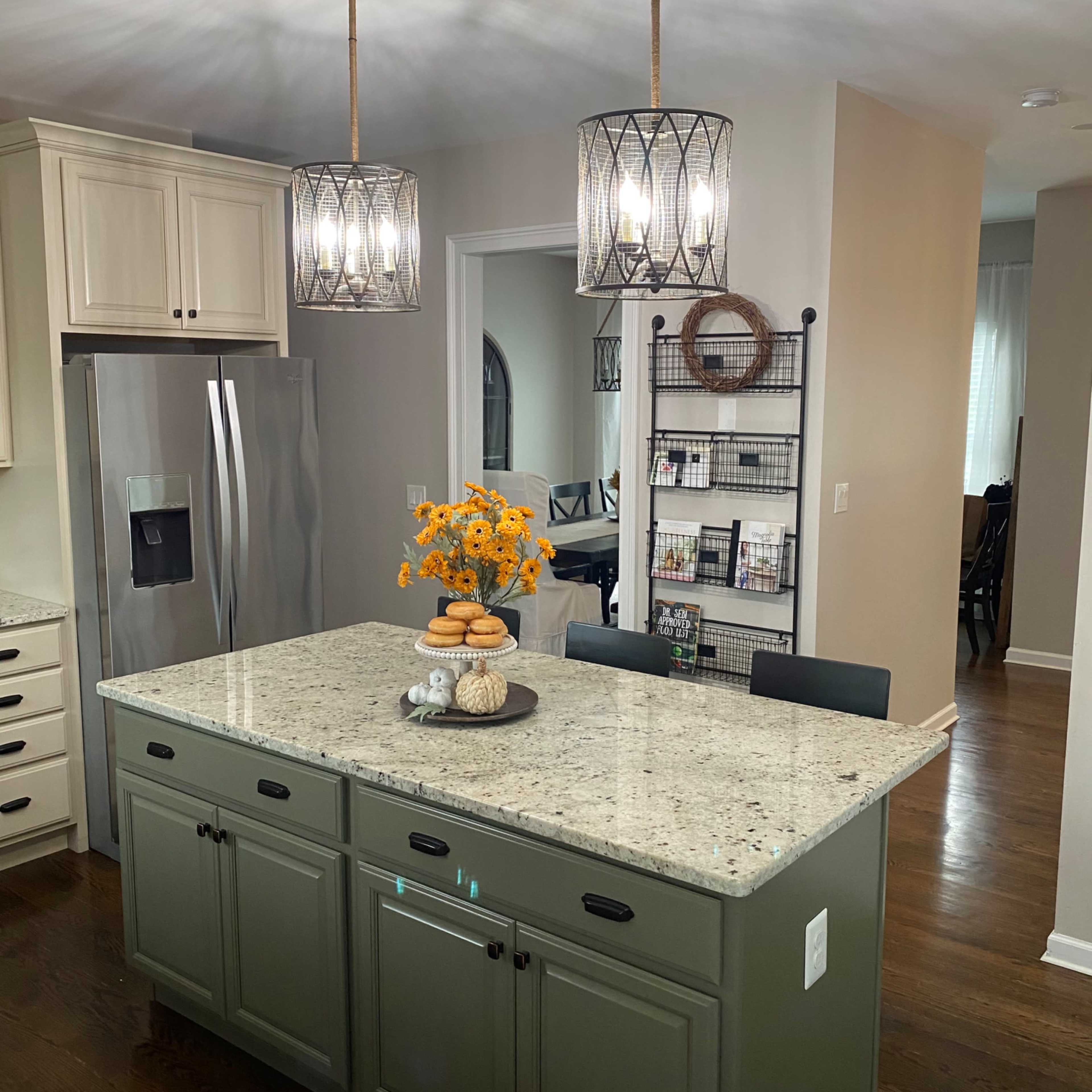 A modern kitchen features a large island with a green base and gray speckled countertop, surrounded by pendant lighting and a dining area in the background.