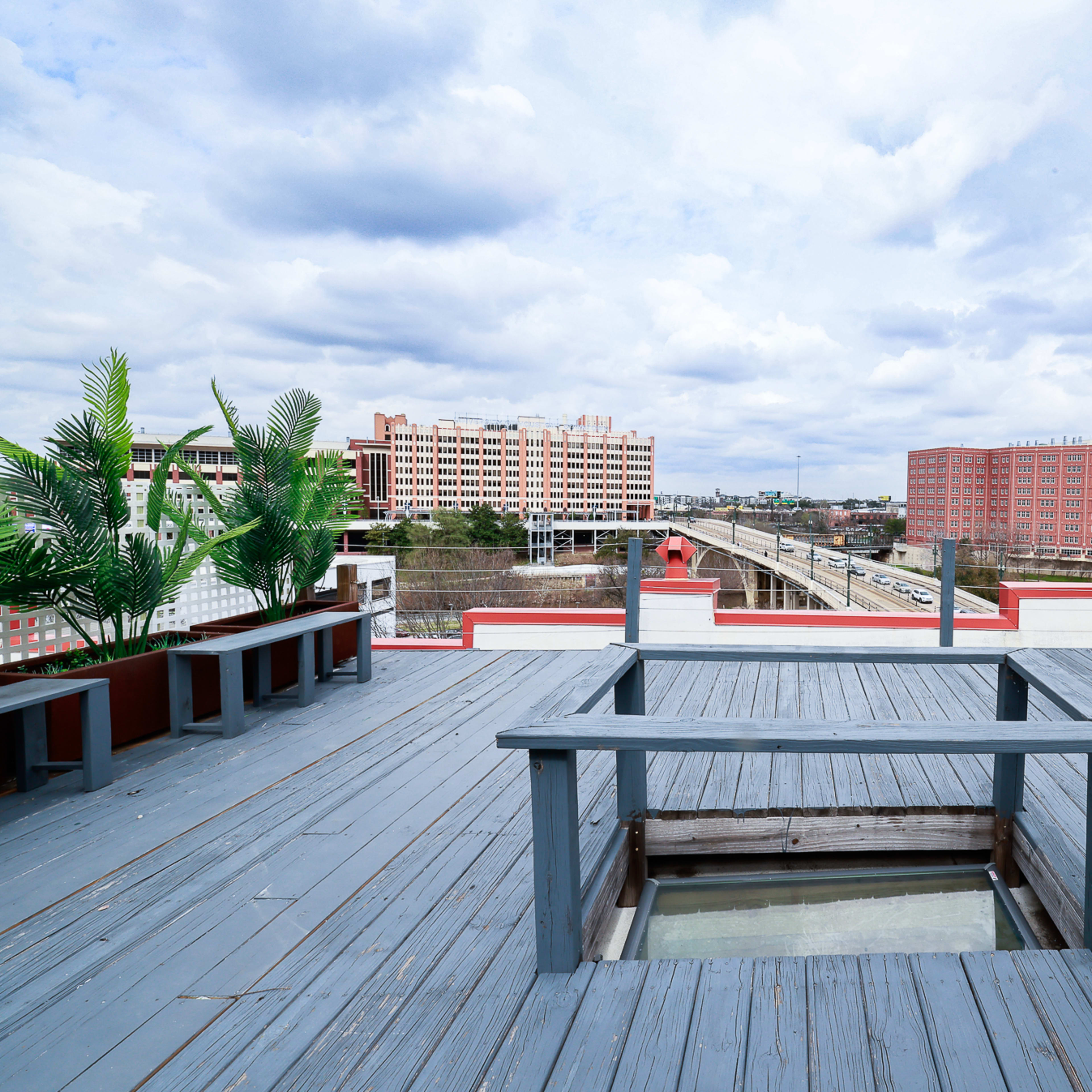 A rooftop deck features wooden flooring and a glass-enclosed area overlooking a highway and city buildings under a cloudy sky.