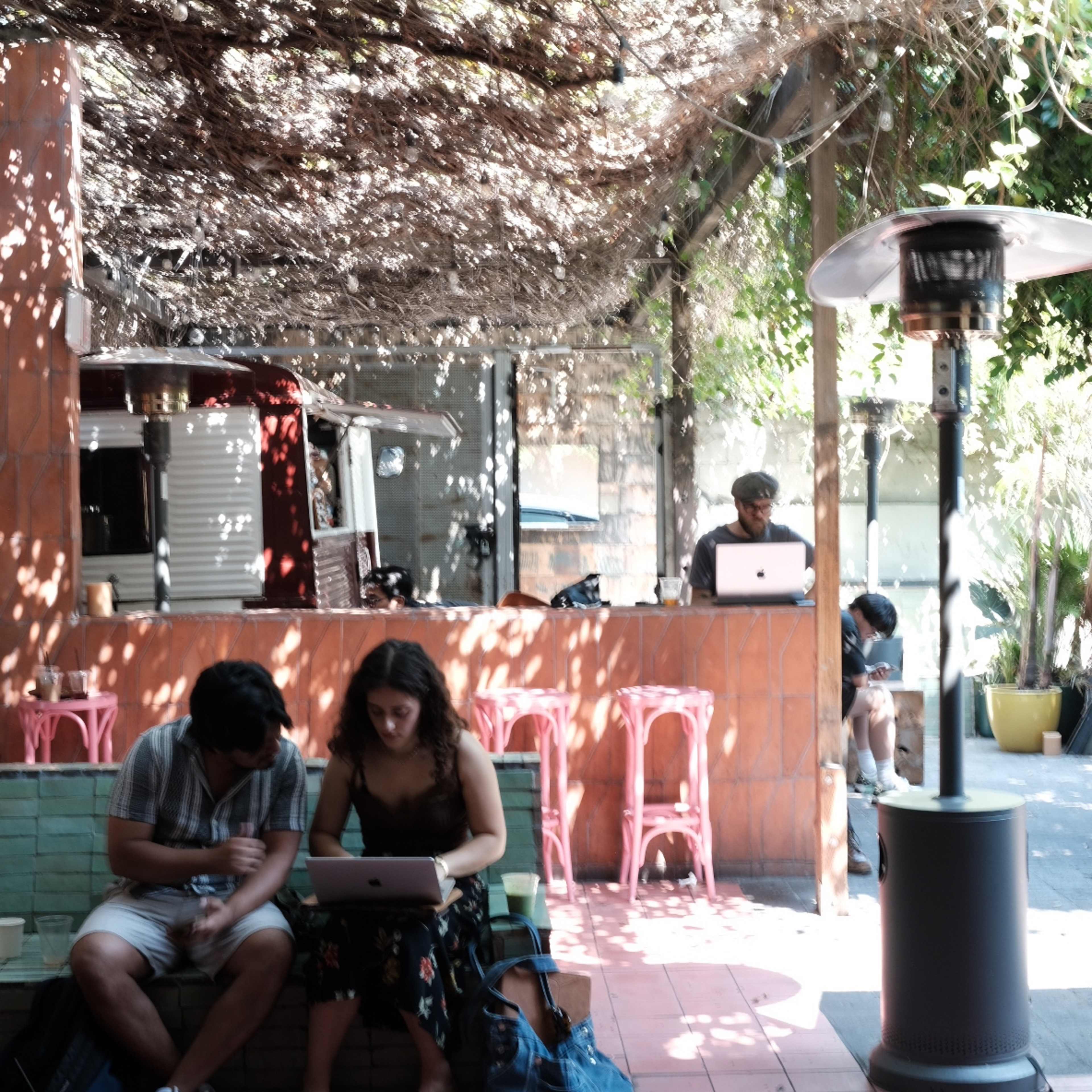 Two people sit on a couch using laptops in a shaded outdoor space with a bar area and a patio heater visible in the background.
