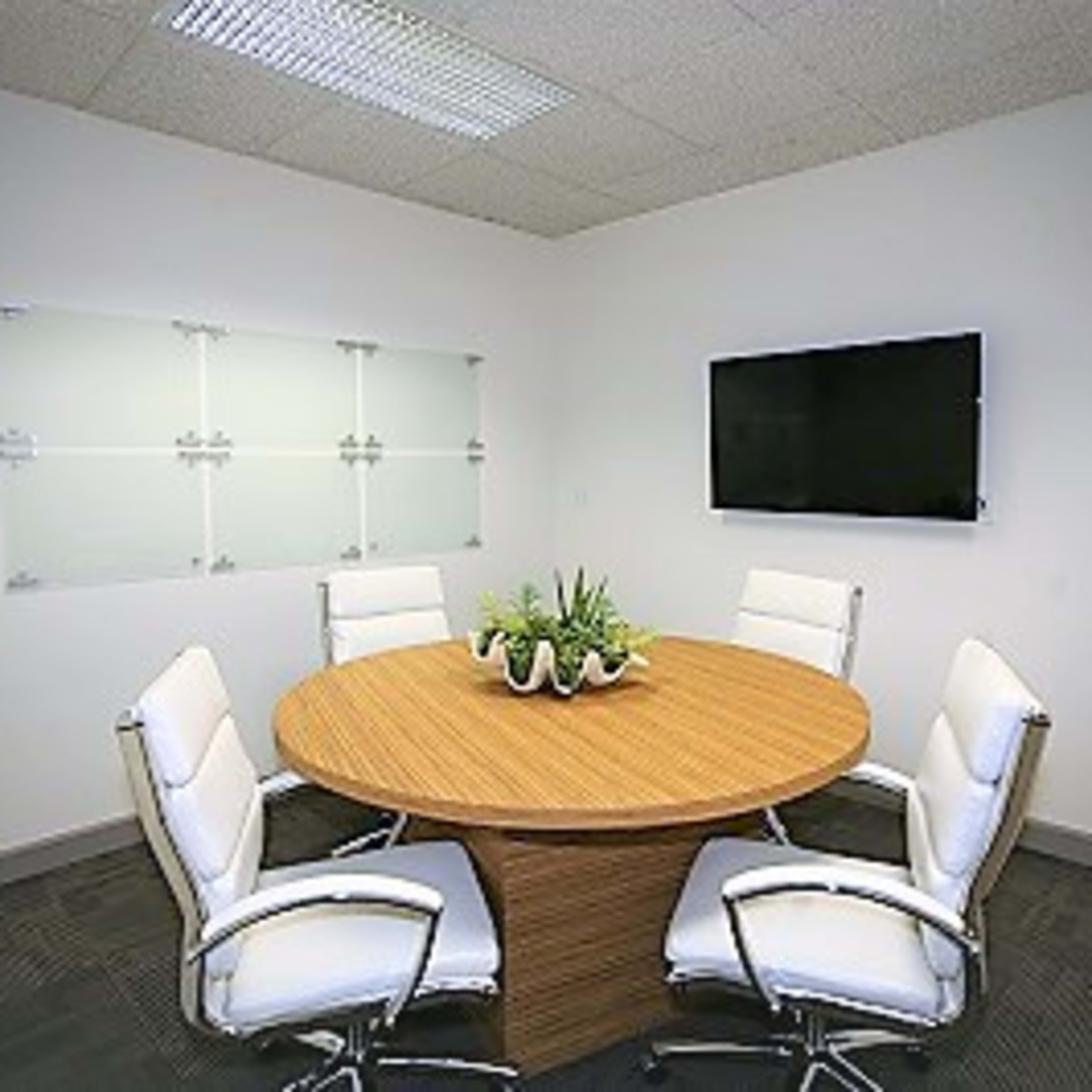 A conference room features a round wooden table surrounded by four white chairs, with a television mounted on the wall and a plant centerpieces on the table.