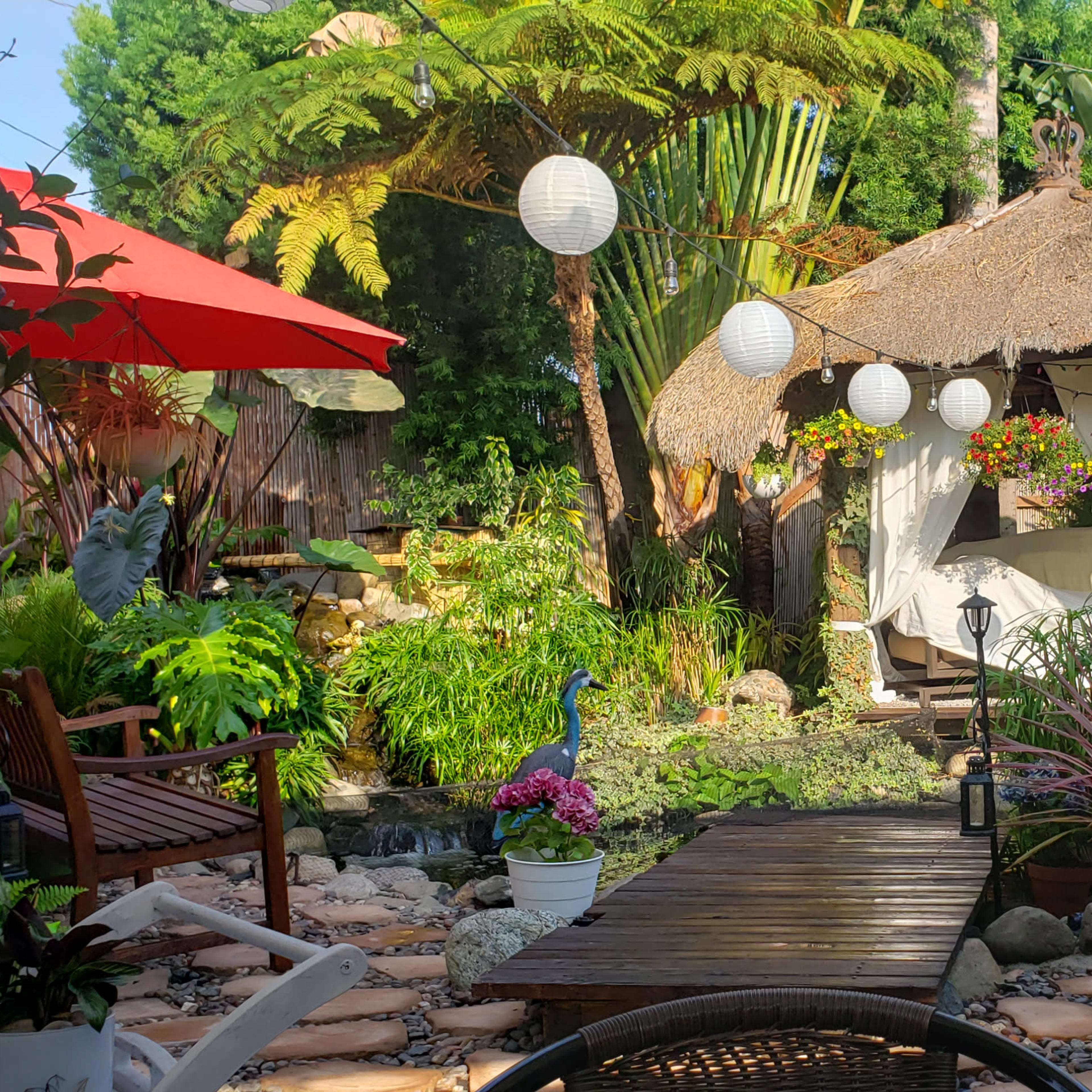 A landscaped garden features a thatched-roof hut surrounded by various plants, flowers, and decorative lanterns, with multiple seating areas and umbrellas.