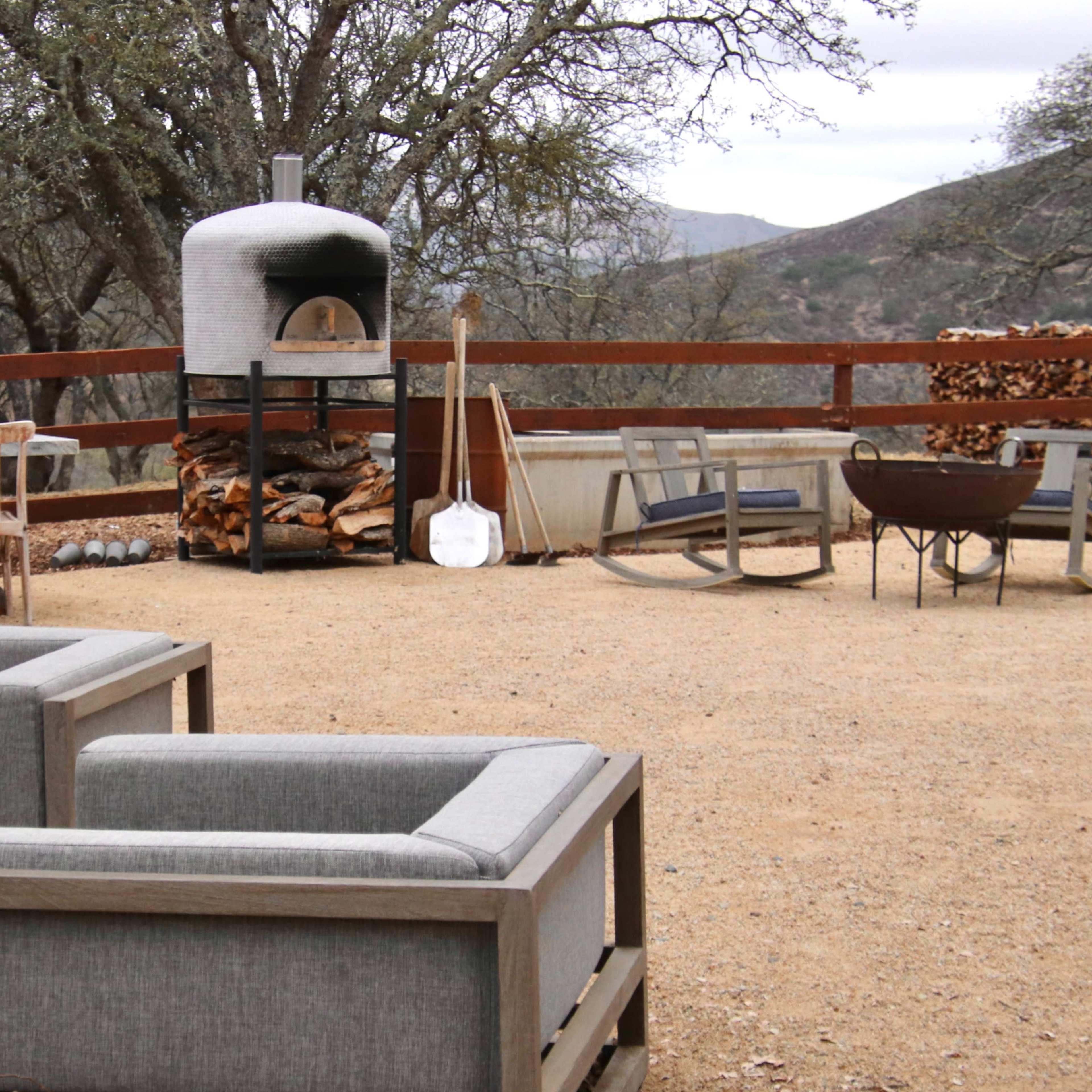A spacious outdoor patio features a stone pizza oven, a wooden dining table, and several comfortable seating arrangements surrounded by trees and a scenic backdrop.