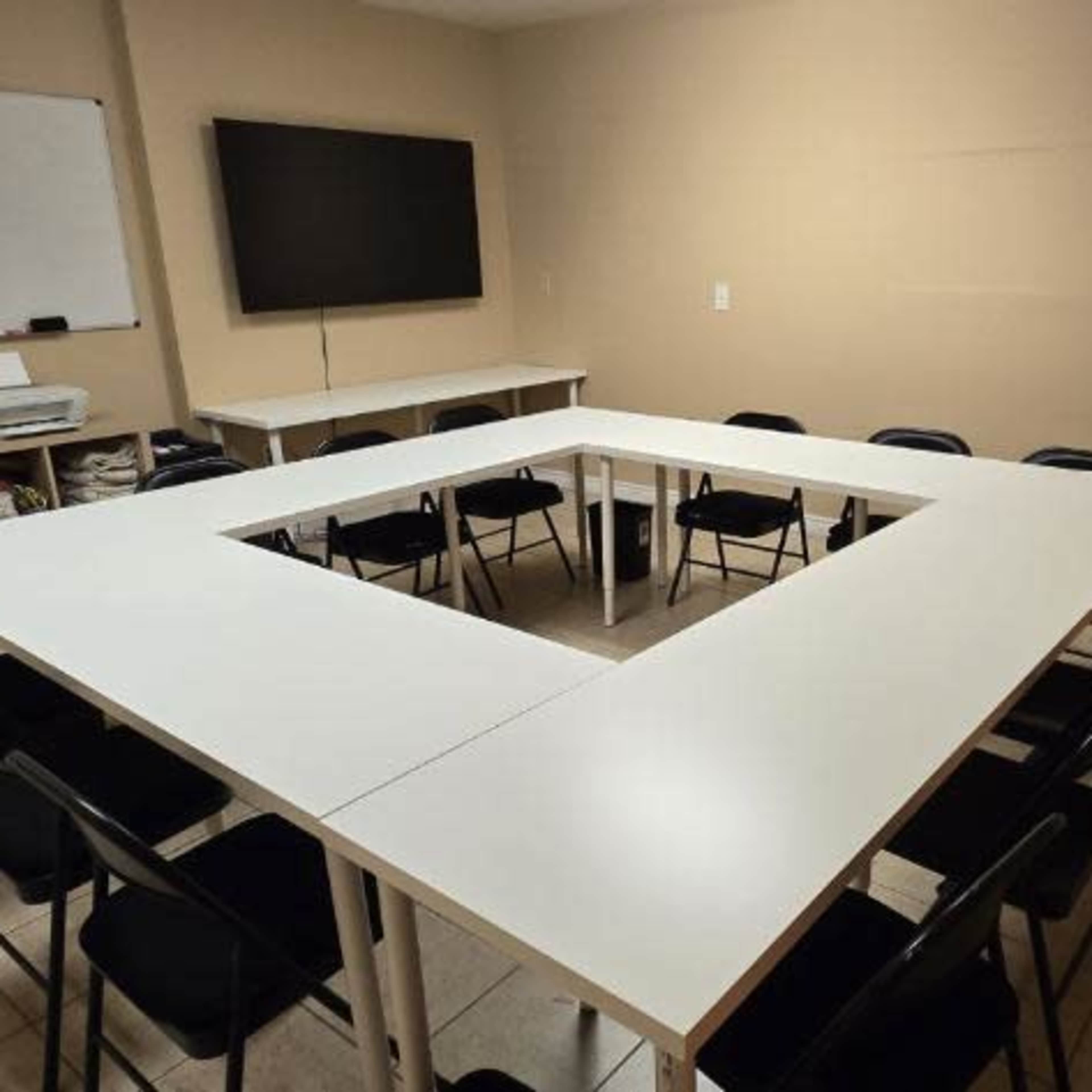 A meeting room features a large square formation of tables surrounded by black chairs, with a whiteboard and a television on the wall.