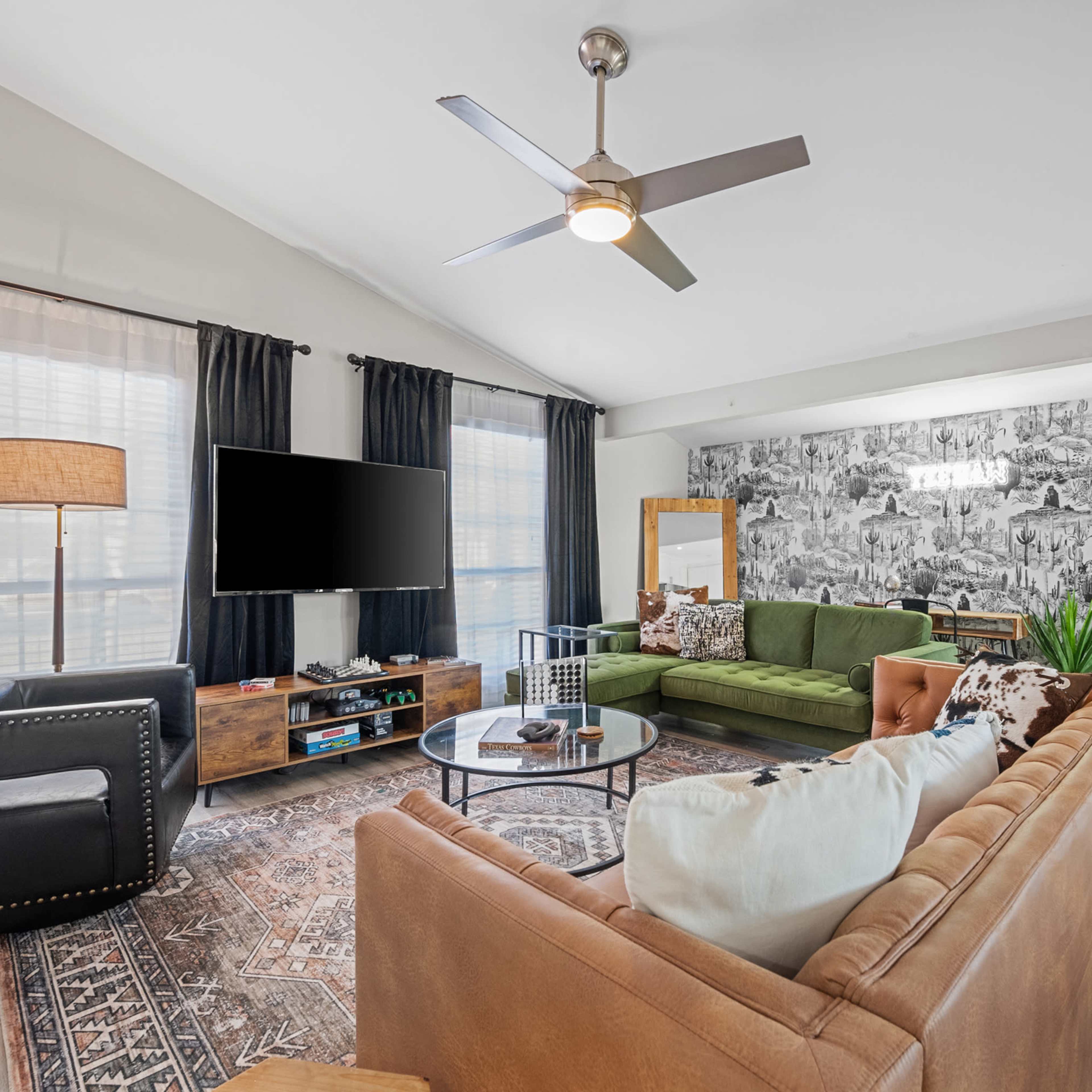 A modern living room features a green sectional sofa, a brown leather couch, a coffee table, and a large wall-mounted television against a patterned wallpaper backdrop.