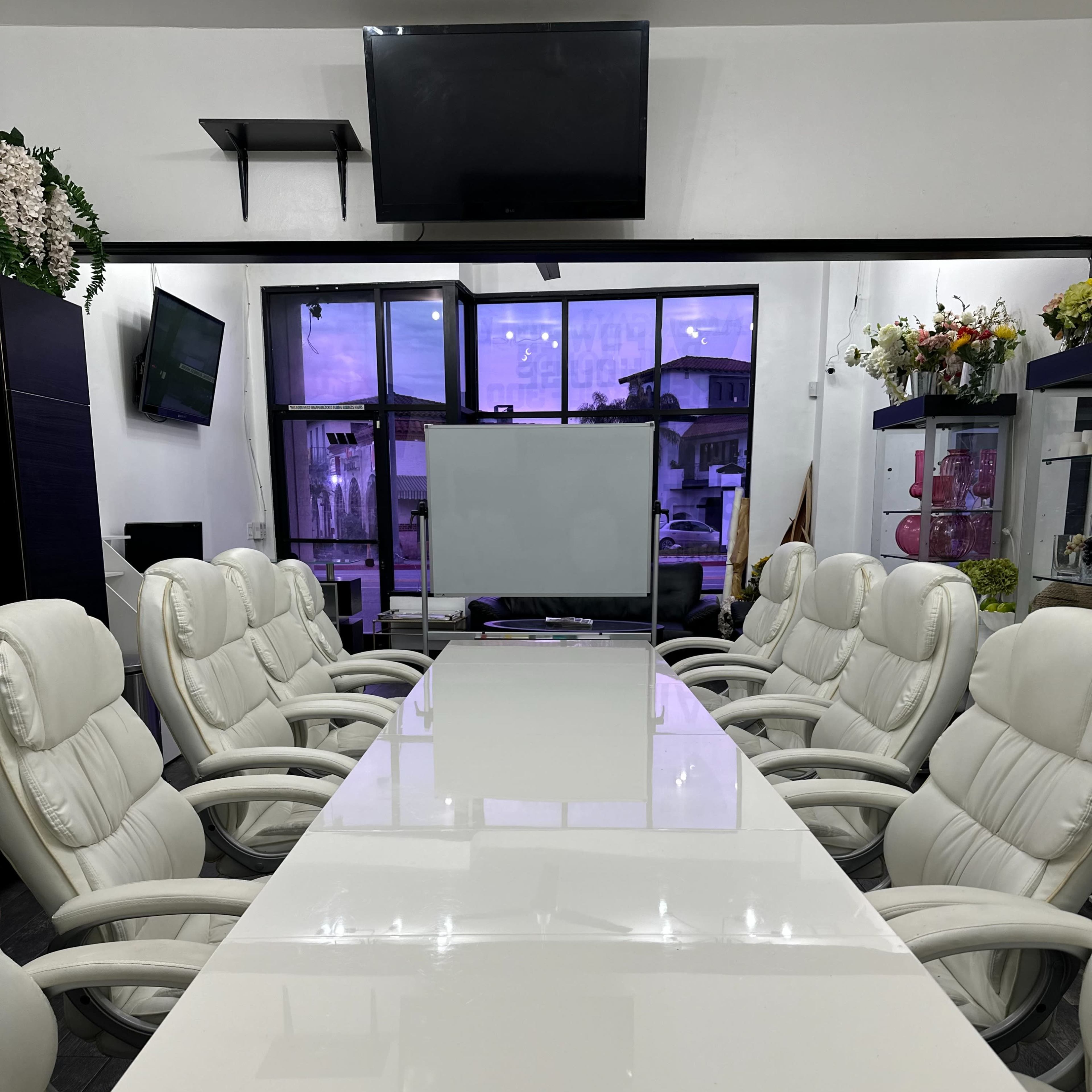 A long, white conference table surrounded by ergonomic chairs is set up in a room with large windows and a wall-mounted television.