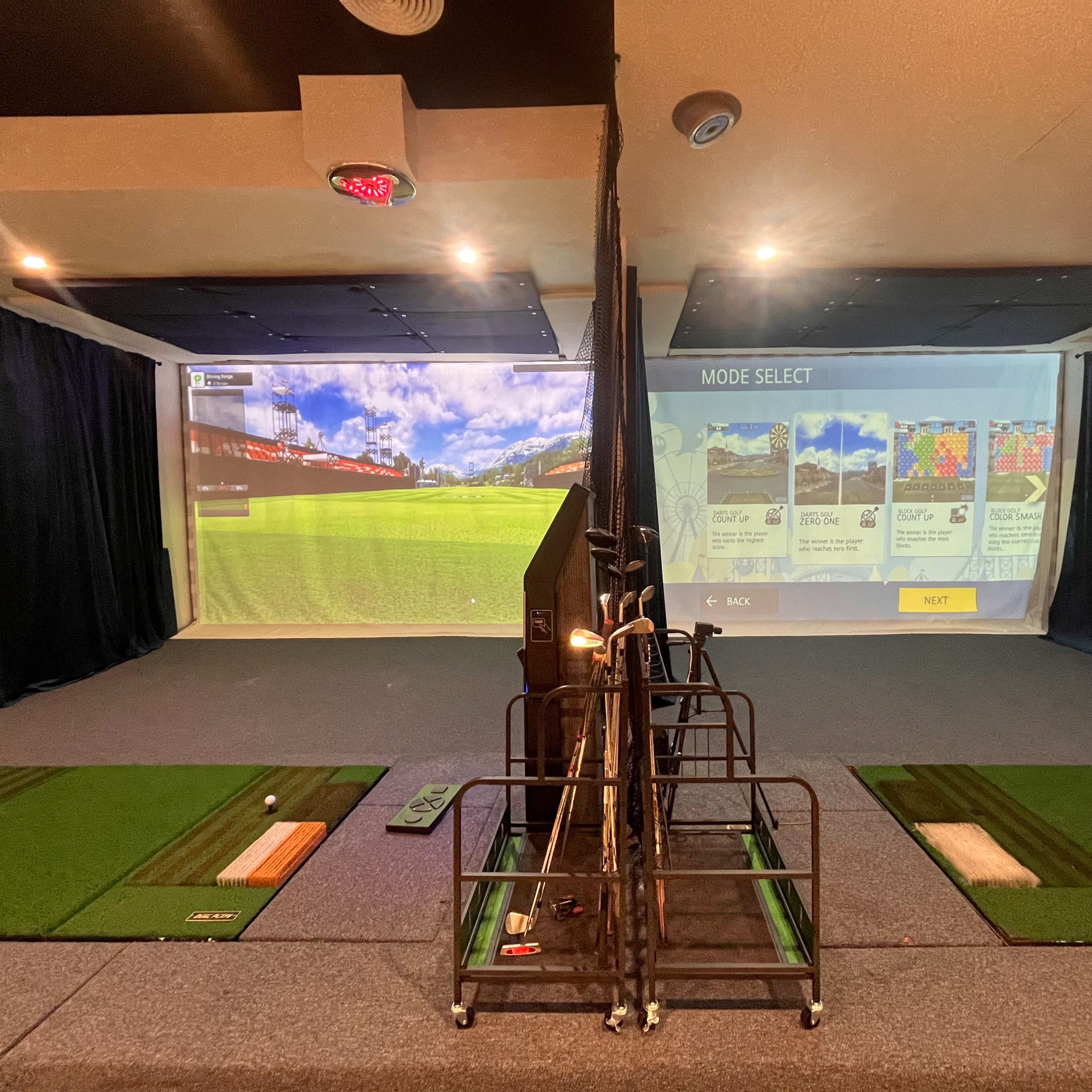 An indoor golf simulation room with artificial grass tee areas and large projection screens displaying golf courses.