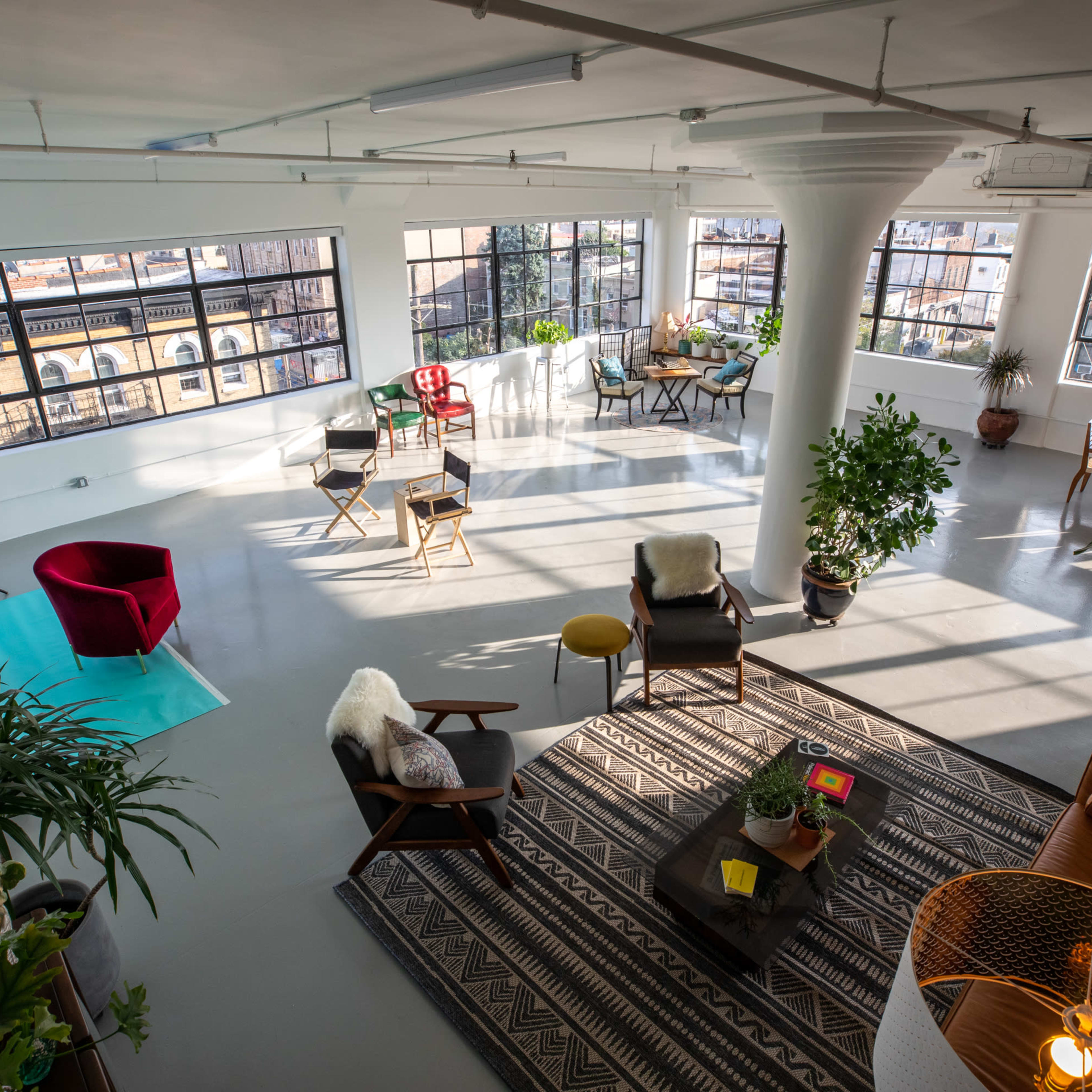 A bright, open studio space with large windows, various seating arrangements, plants, and a vibrant area for activities.