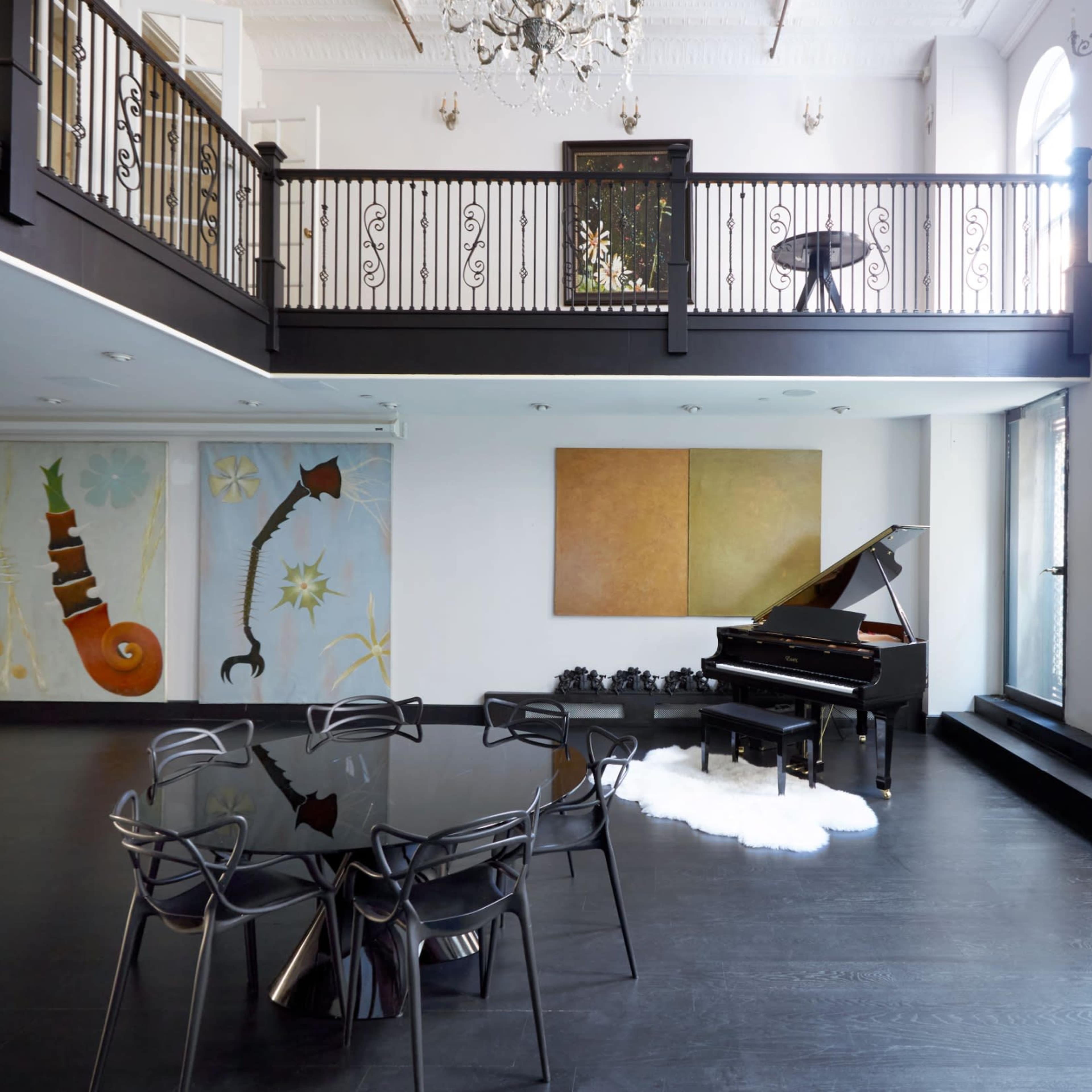 A spacious room with high ceilings, featuring a black dining table surrounded by transparent chairs, a grand piano, and large windows with modern artwork on the walls.