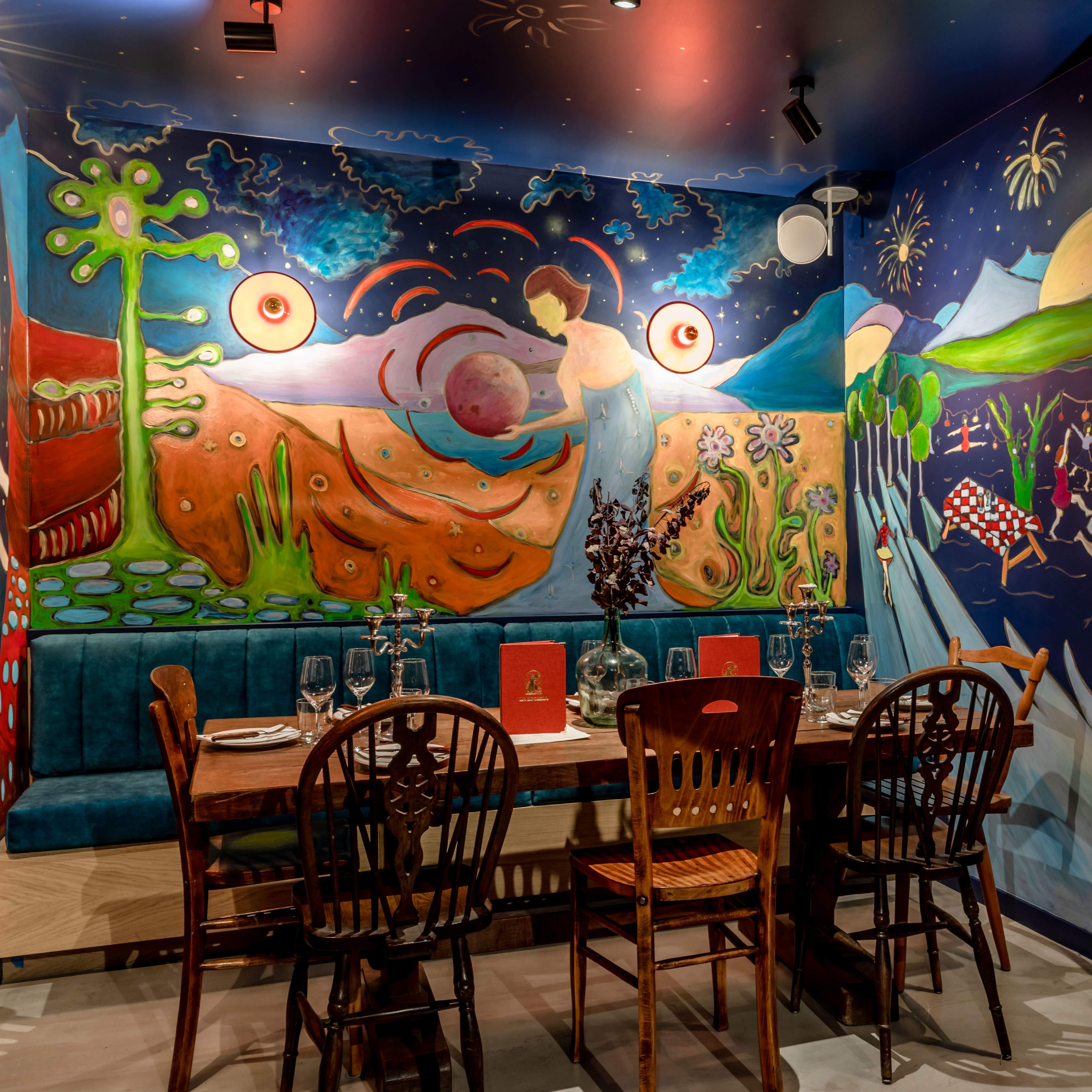 A vibrant dining area with a large, colorful mural on the walls, featuring a variety of abstract shapes and designs, and a wooden table set for a meal.