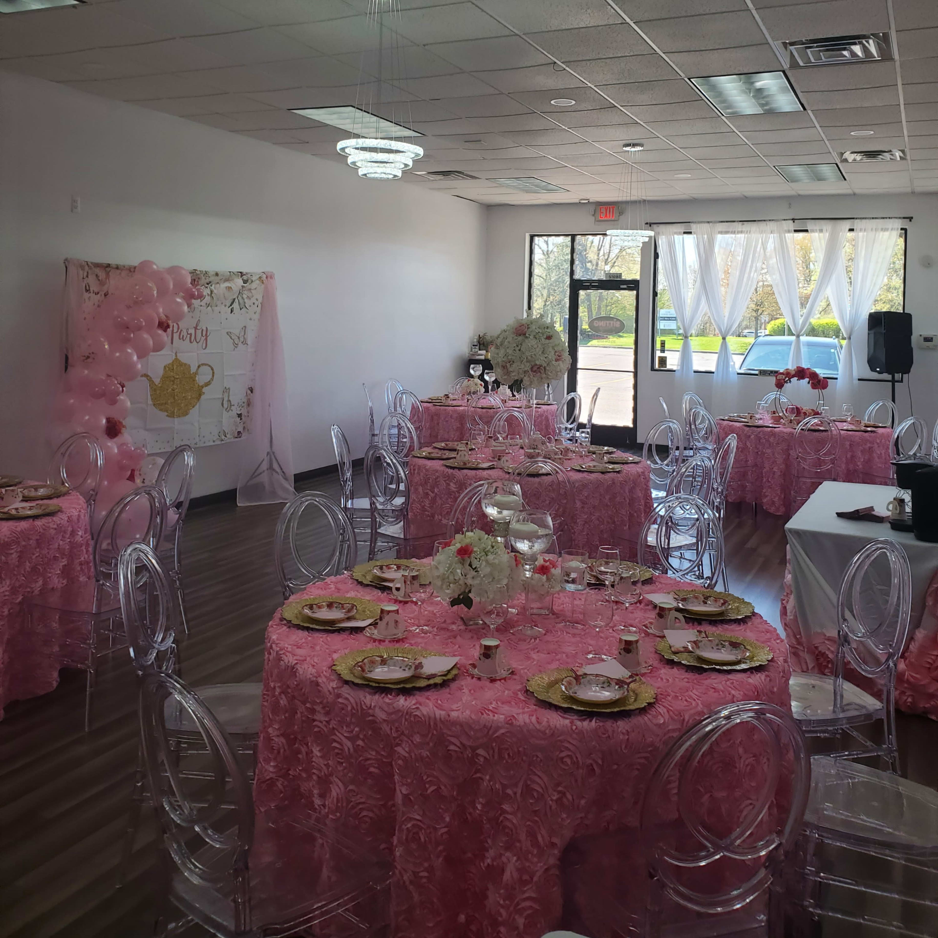 A brightly lit event space decorated with pink tablecloths, clear chairs, and floral centerpieces, set up for a formal gathering.