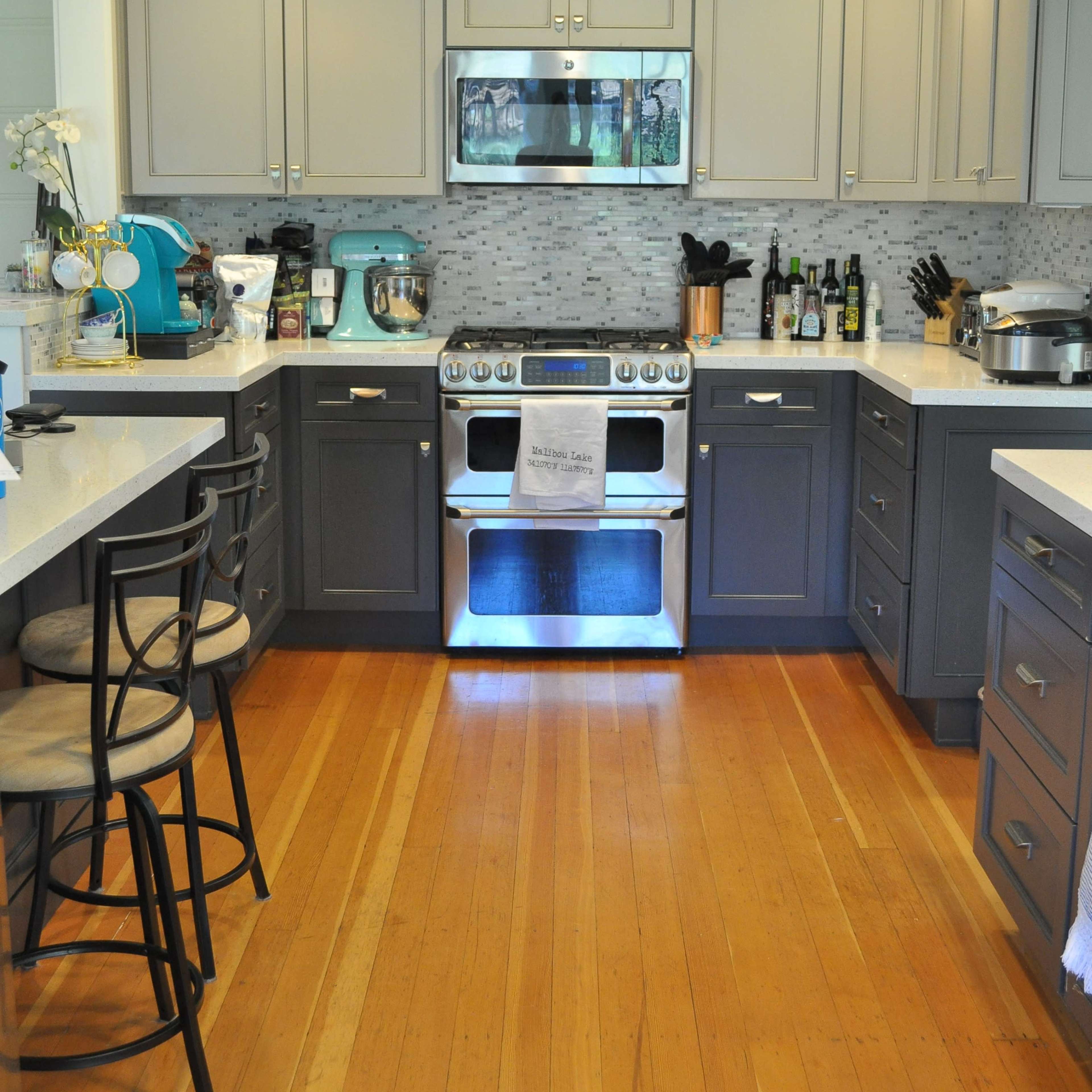 A modern kitchen features gray cabinets, a stainless steel stove, and a wooden floor with a breakfast bar and seating.