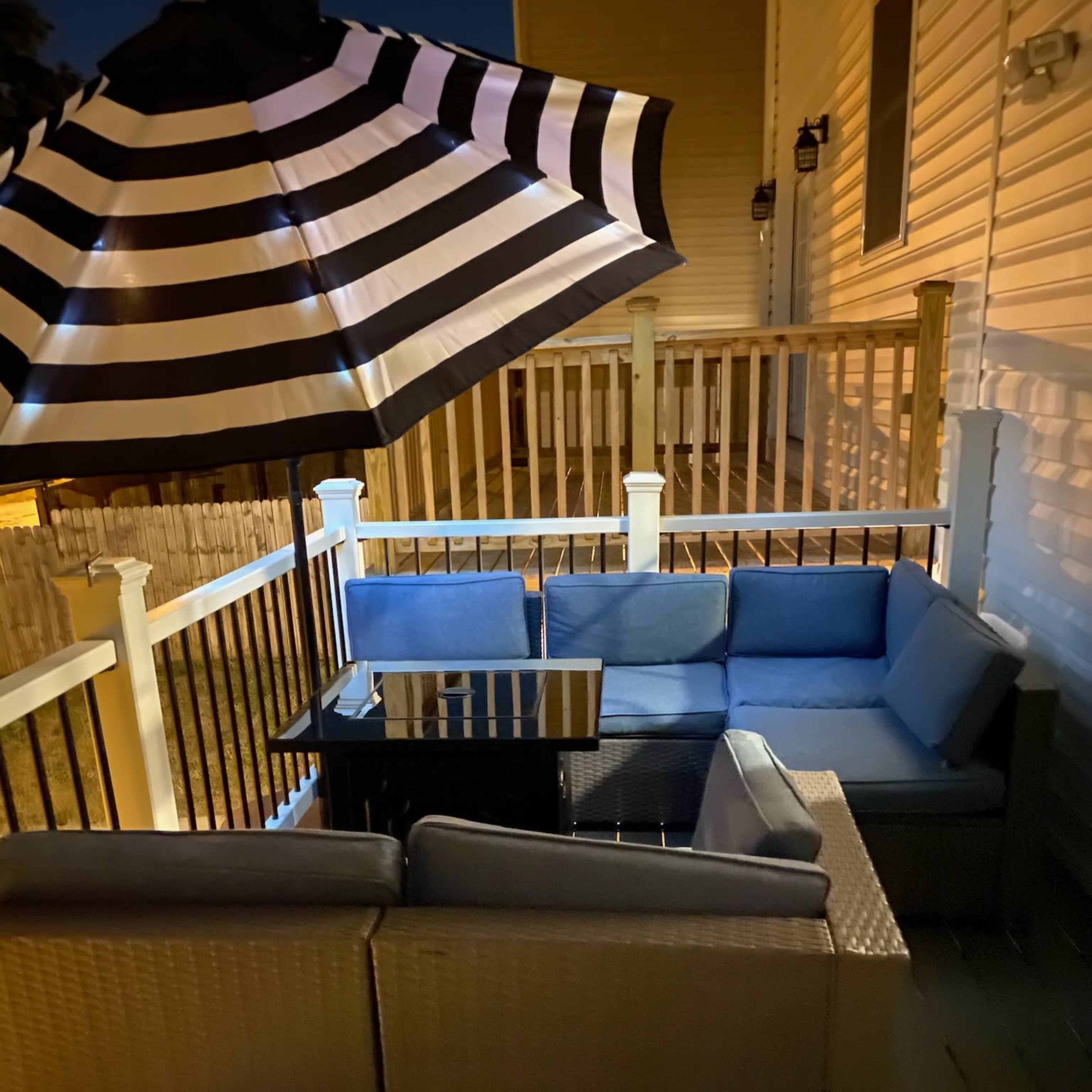 A patio seating area features a dark table surrounded by blue cushions and a striped umbrella above it, all situated on a wooden deck at night.