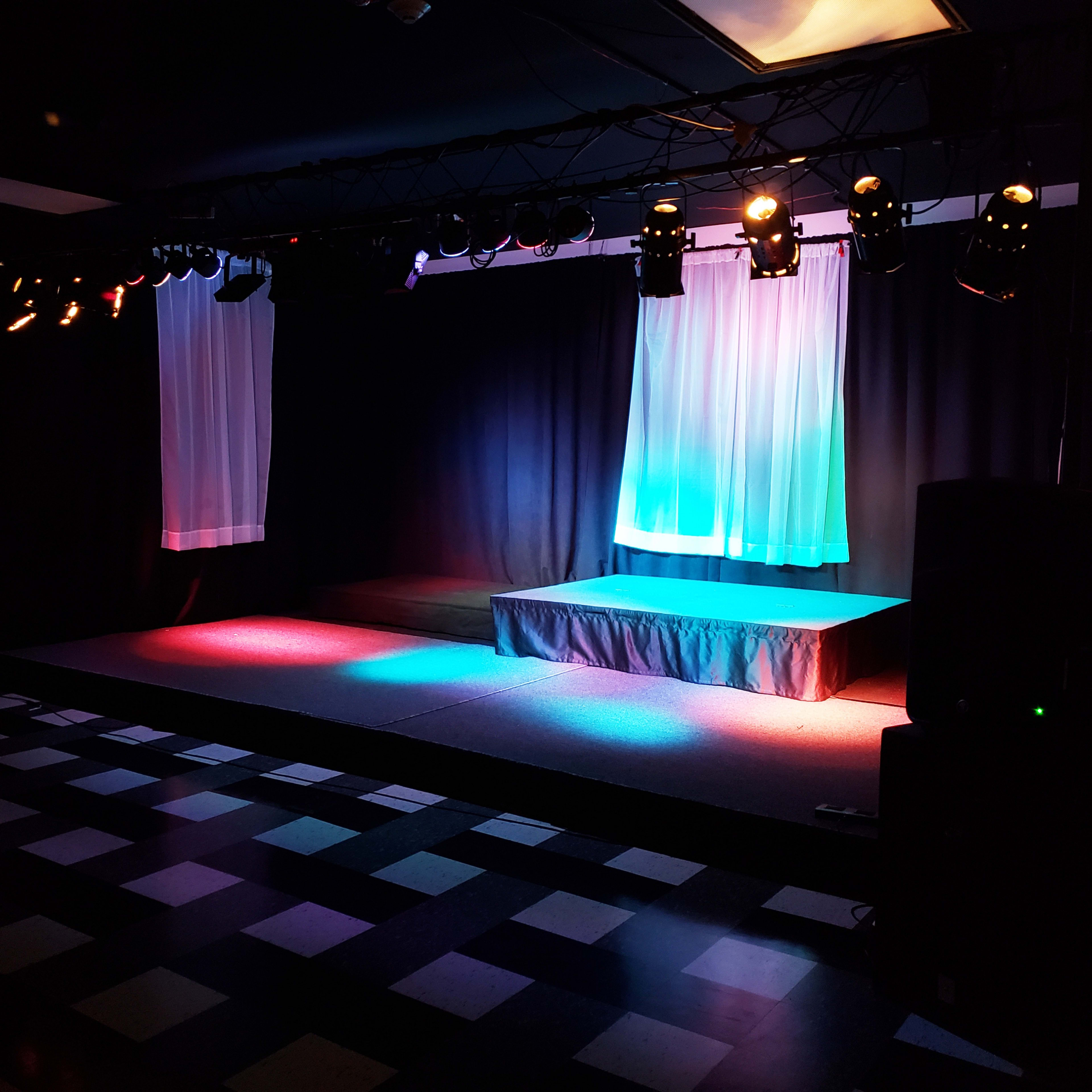 A stage with colored lighting, draped fabric in the background, and speakers on either side, set against a checkered floor.