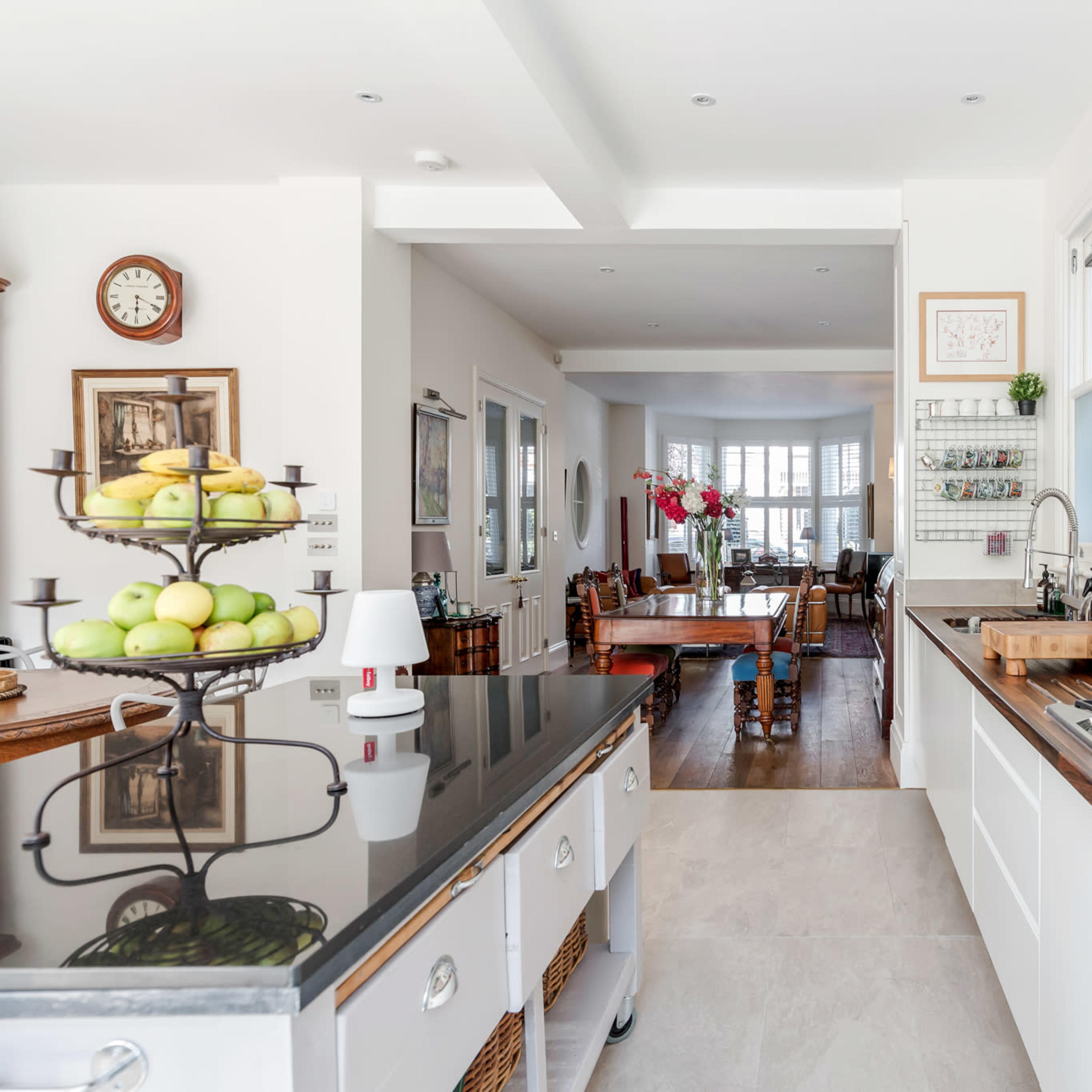 A modern kitchen features a central island with a fruit display, stainless steel appliances, and a view into a dining area with a wooden table.