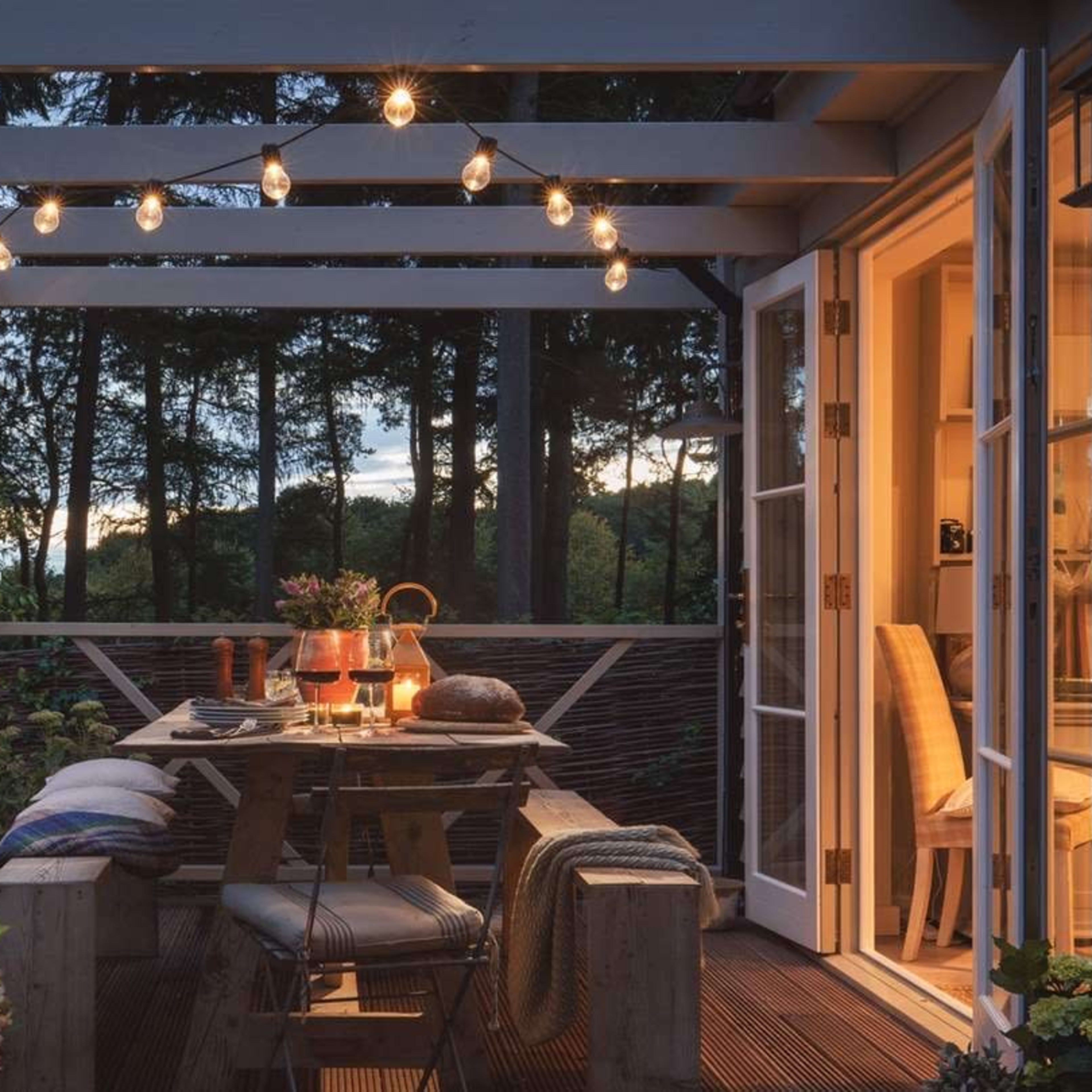 A wooden patio with string lights above a dining table set for a meal, surrounded by greenery and trees at dusk.