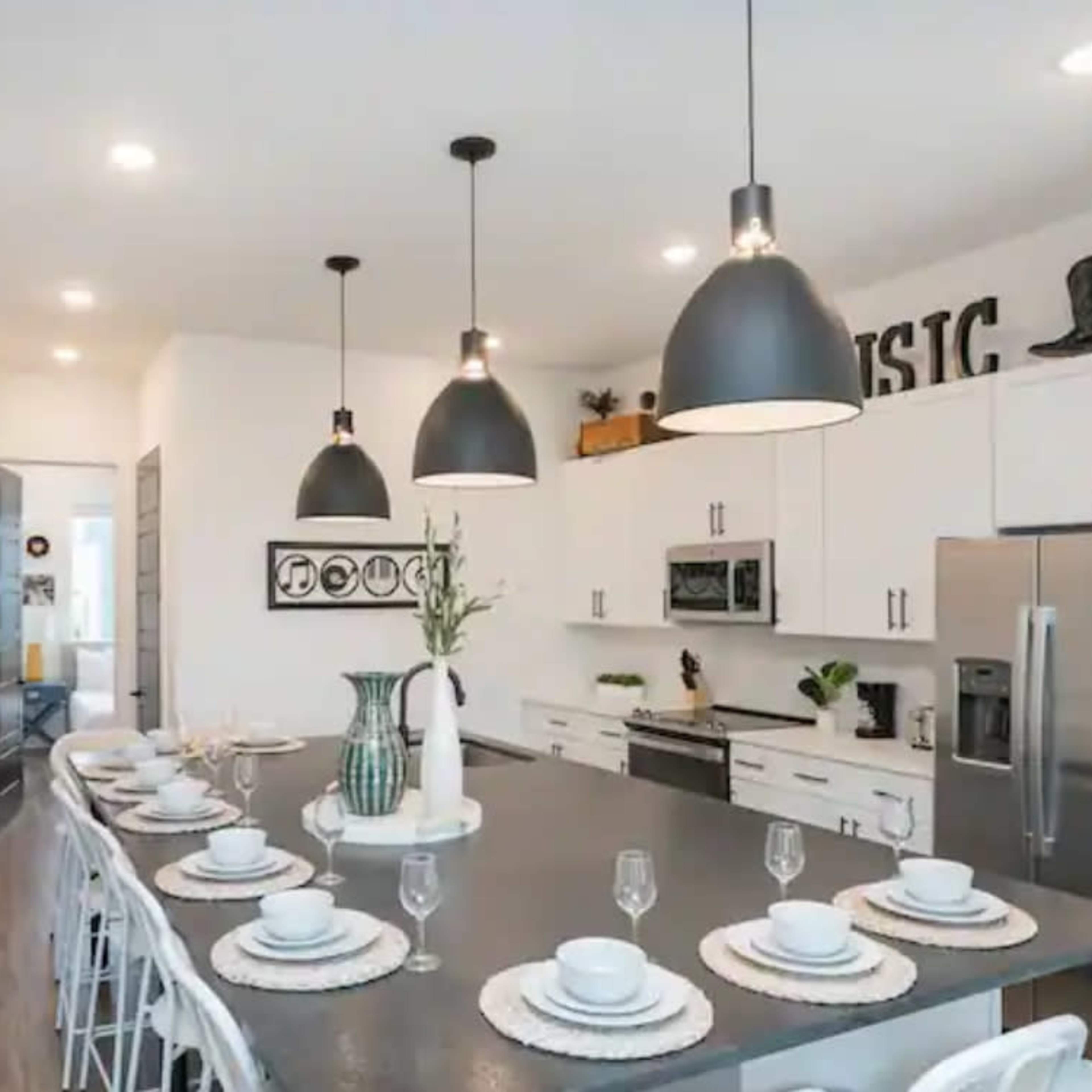 A modern kitchen features a long island with dining settings, pendant lights, and contemporary appliances.