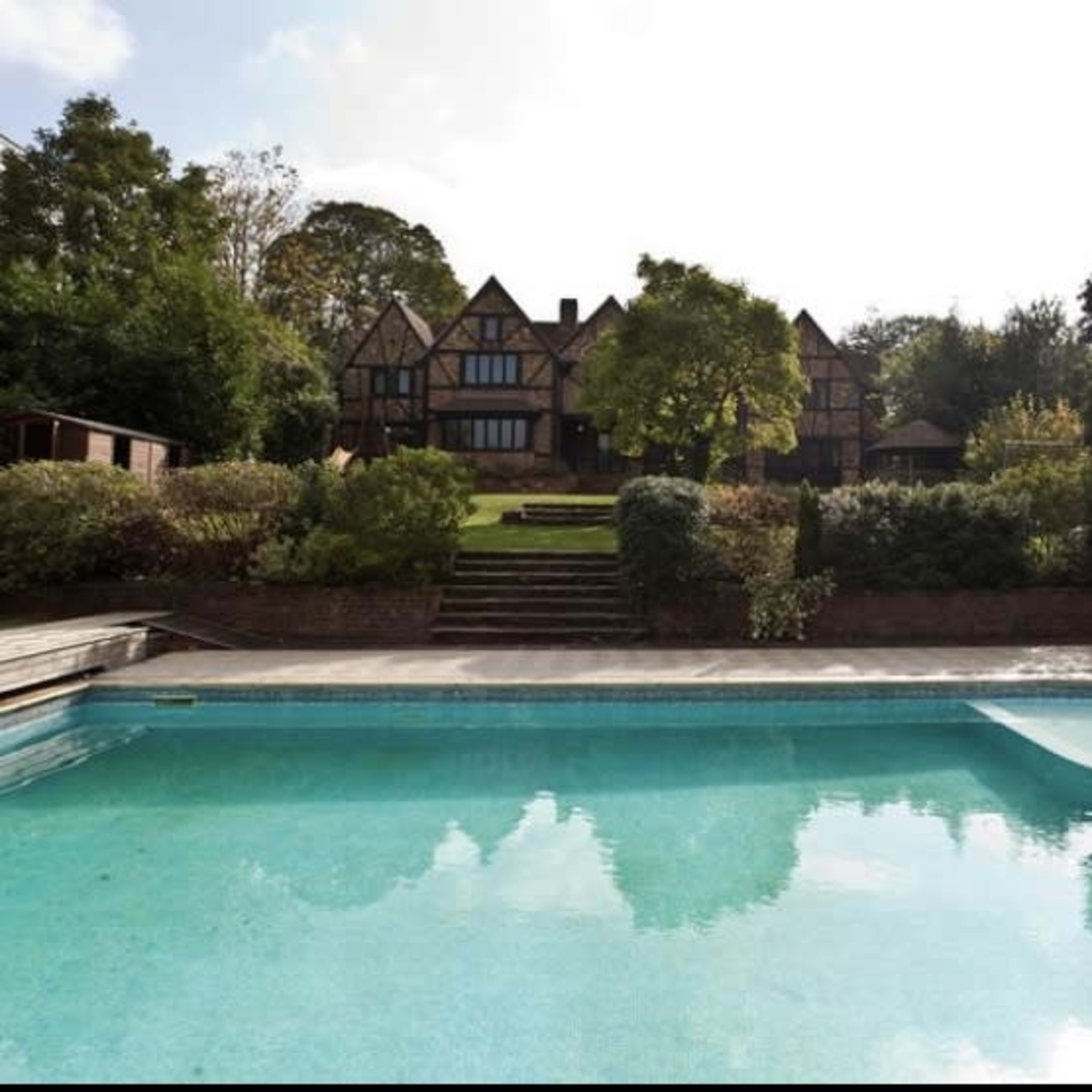A large, traditional-style house is visible in the background, set on a hillside next to a swimming pool surrounded by landscaped greenery.