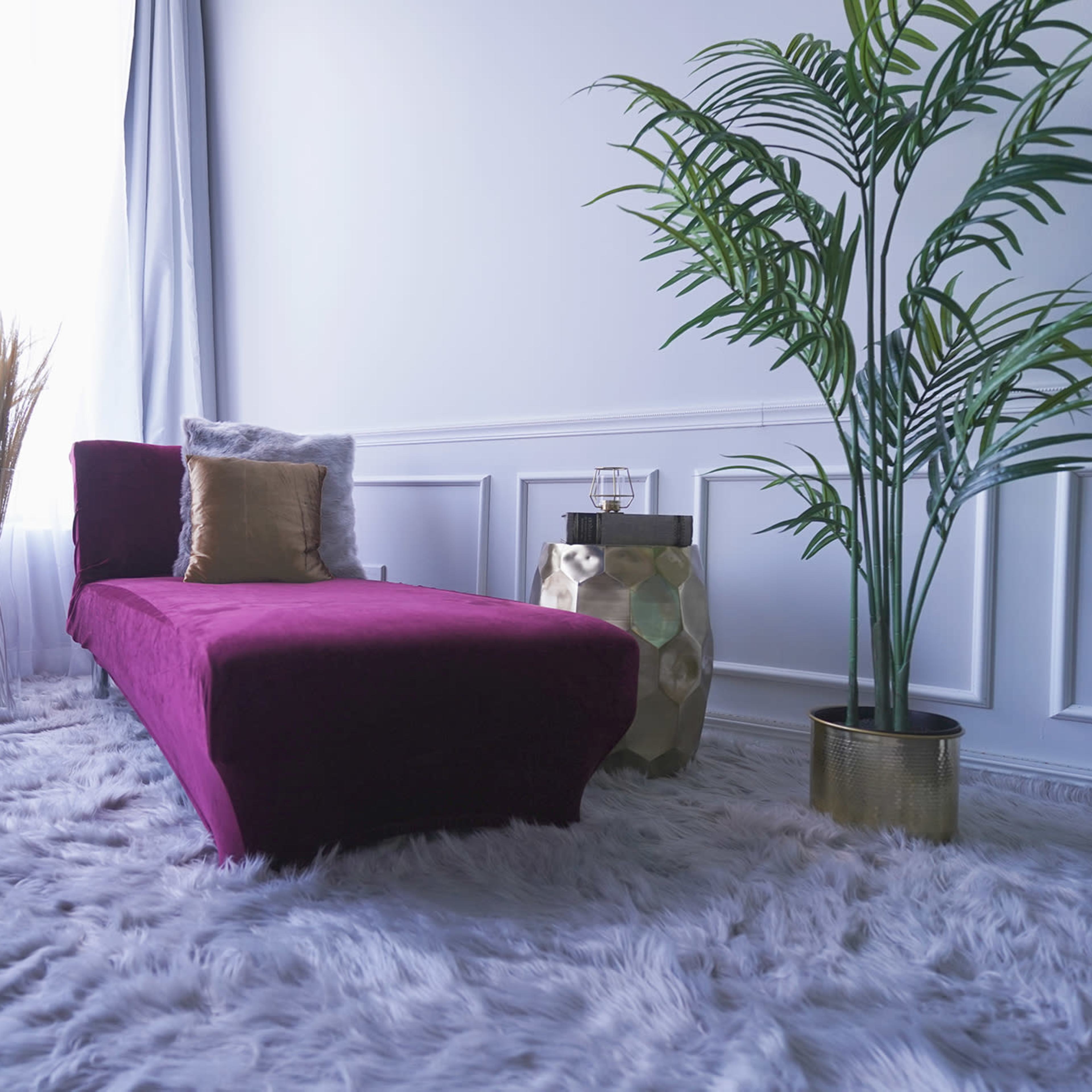 A purple chaise lounge placed on a plush, white area rug, next to a potted plant and a small side table with glassware.