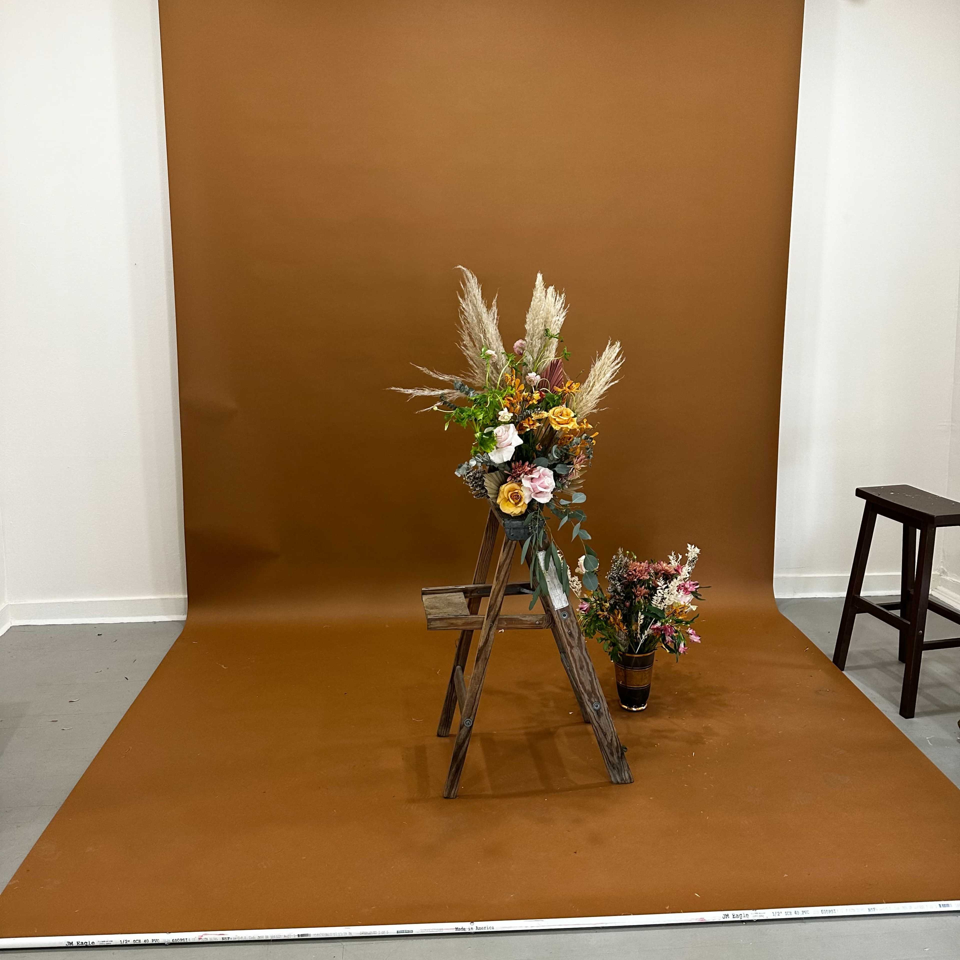 A photography studio setup featuring a brown backdrop, a wooden ladder display with floral arrangements, and a potted plant on the floor.