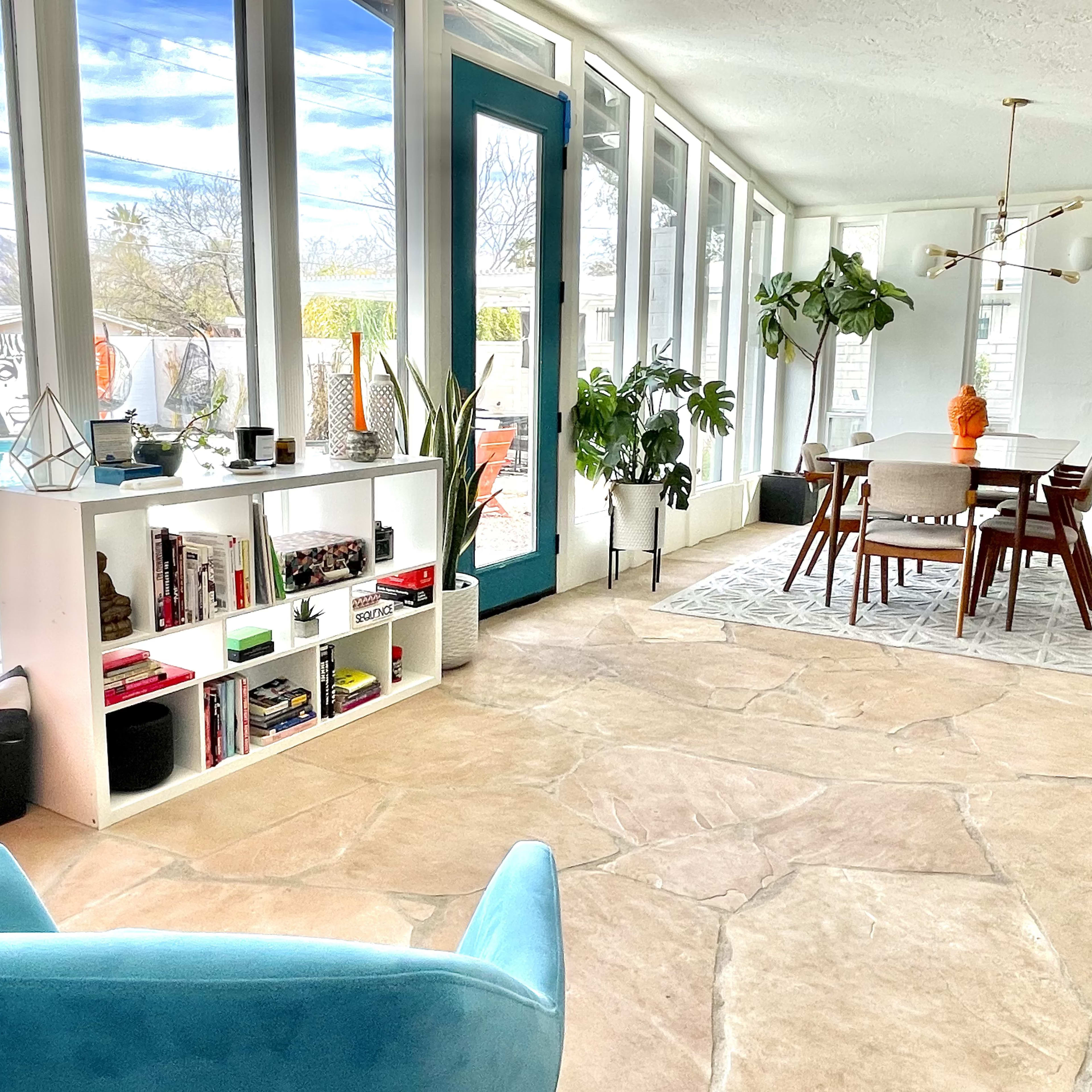 A bright, modern living space with a dining area, a bookshelf, large windows, and a view of an outdoor pool.