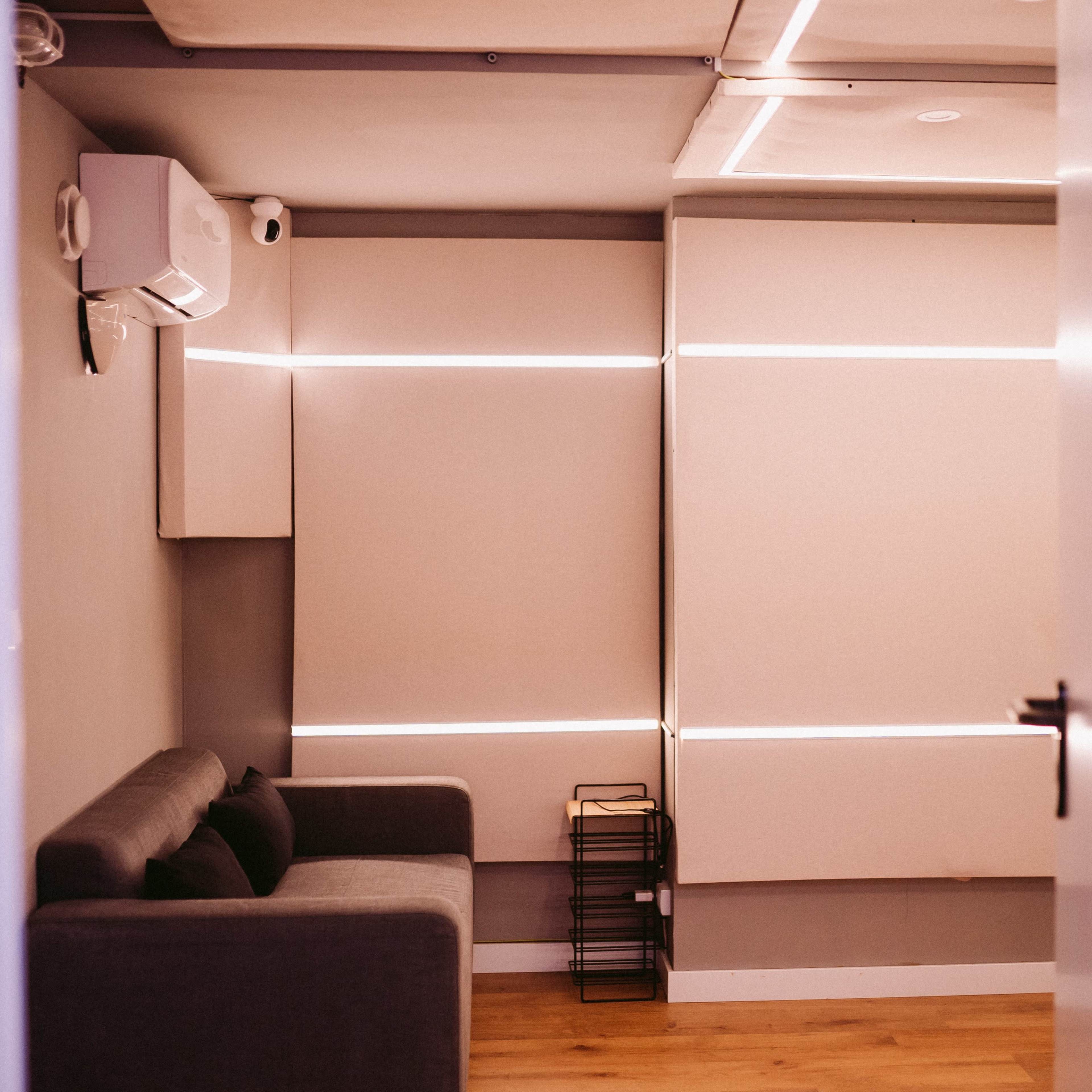 A small, modern room features a gray sofa, wall-mounted air conditioning unit, and illuminated wall panels.