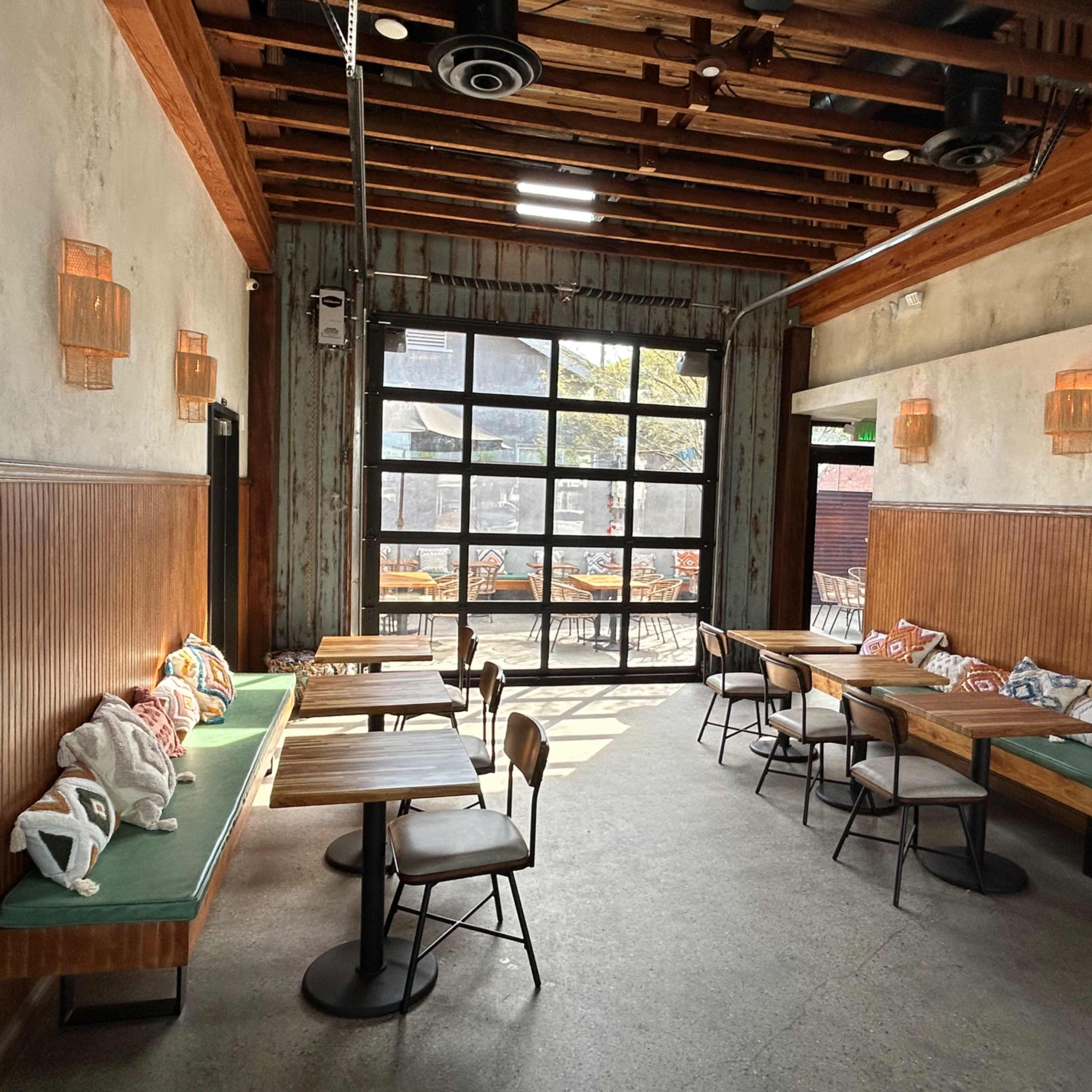 The interior of a restaurant features wooden beams, a large glass door, and minimally furnished tables with patterned cushions.