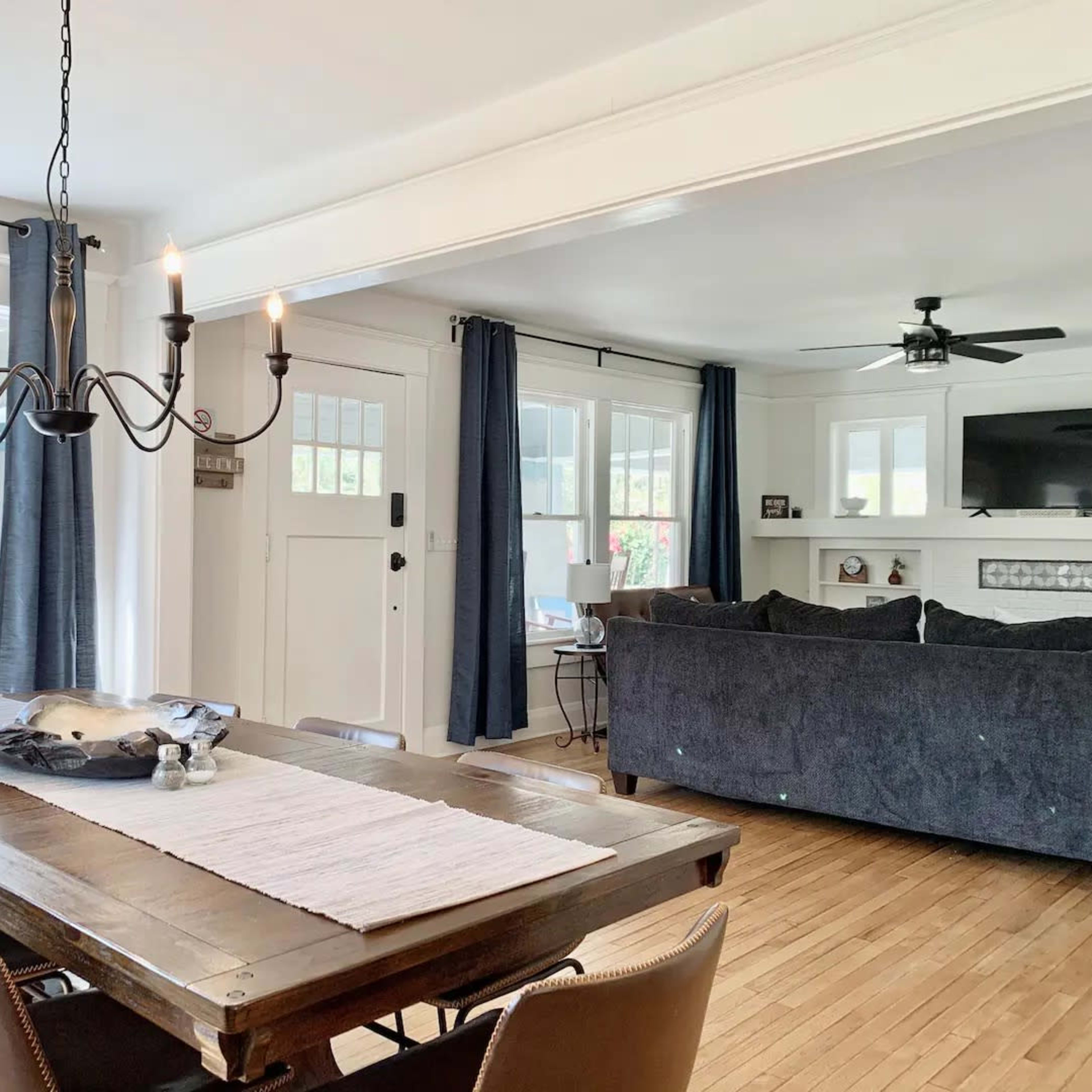 A dining area features a wooden table with chairs, a chandelier overhead, and an open view into a living space with a couch and television.