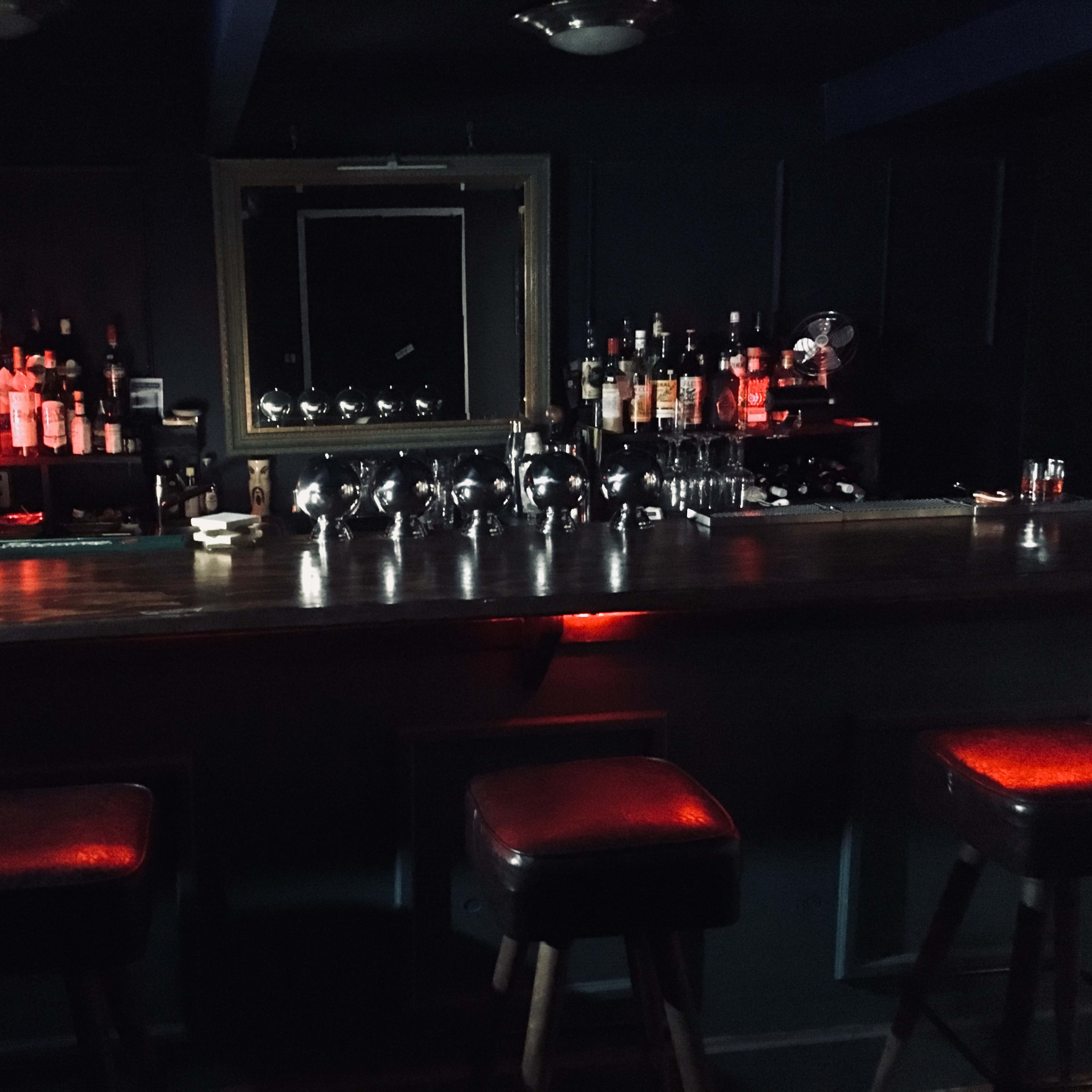 A dimly lit bar with a mirrored back, various bottles displayed on shelves, and three stools against the counter.