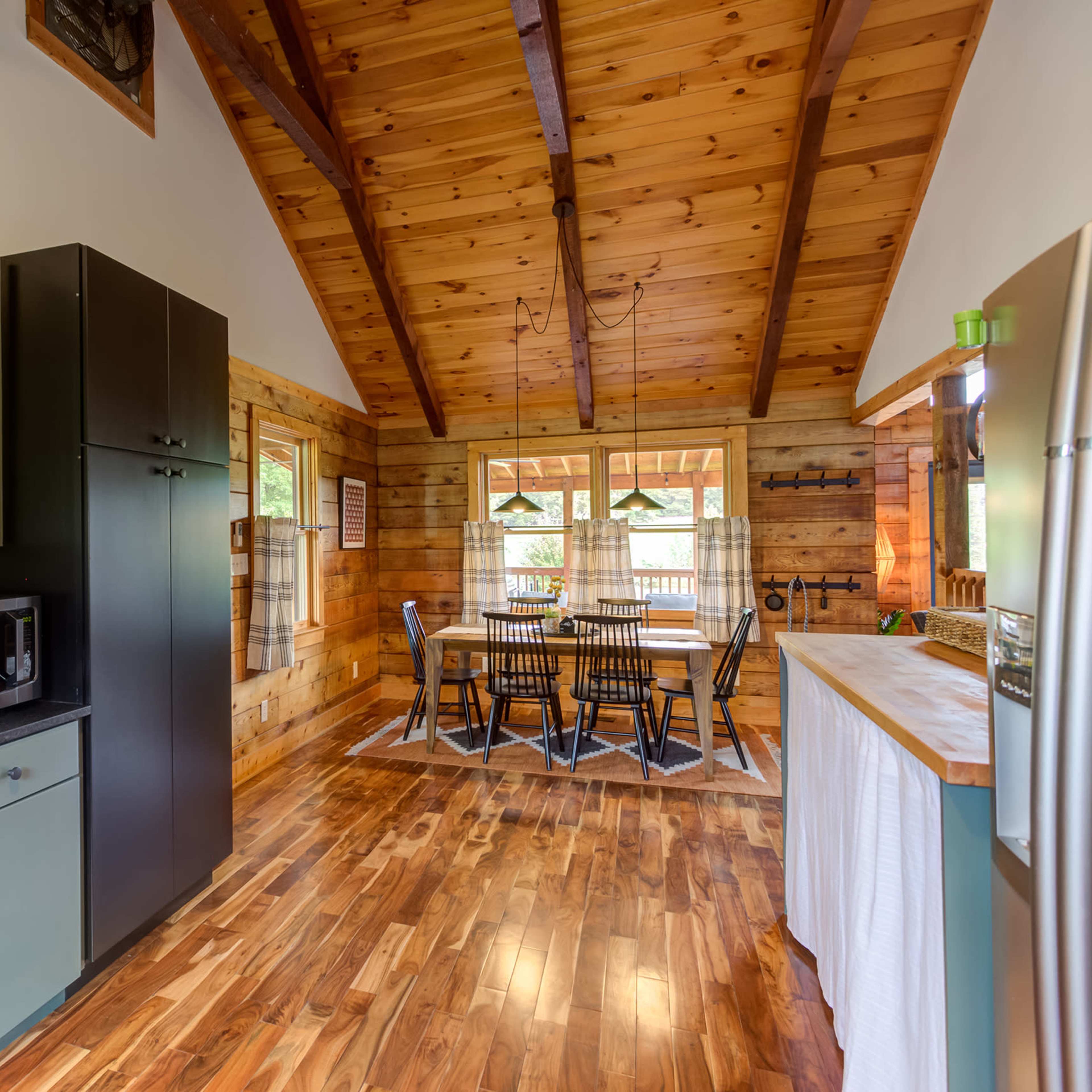 A modern kitchen with green cabinetry, a black refrigerator, and a wooden dining area featuring a table and chairs under a high, exposed beam ceiling.